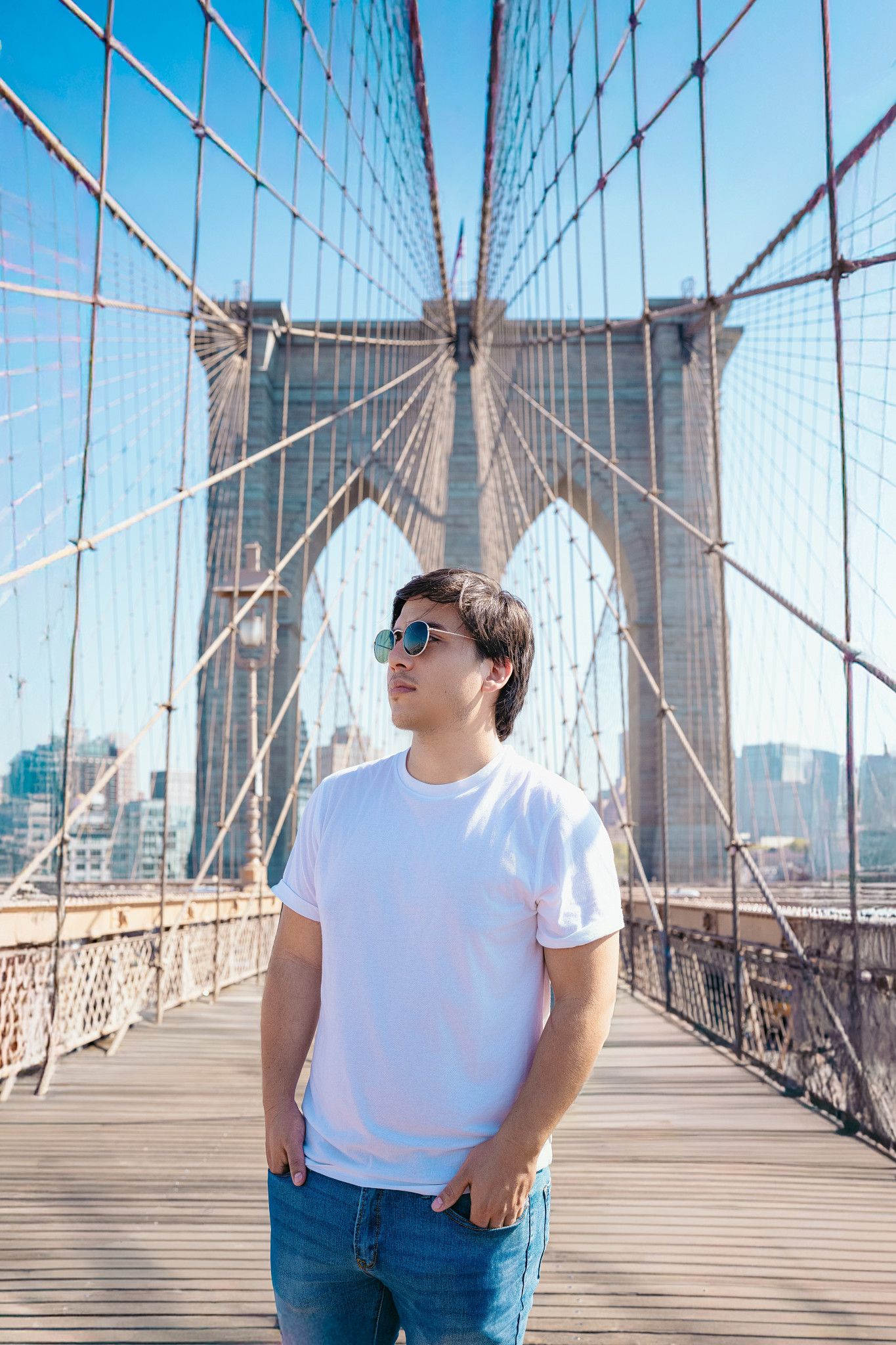 Young man in white t-shirt and sunglasses standing on Brooklyn Bridge walkway with iconic stone tower and cables behind him.