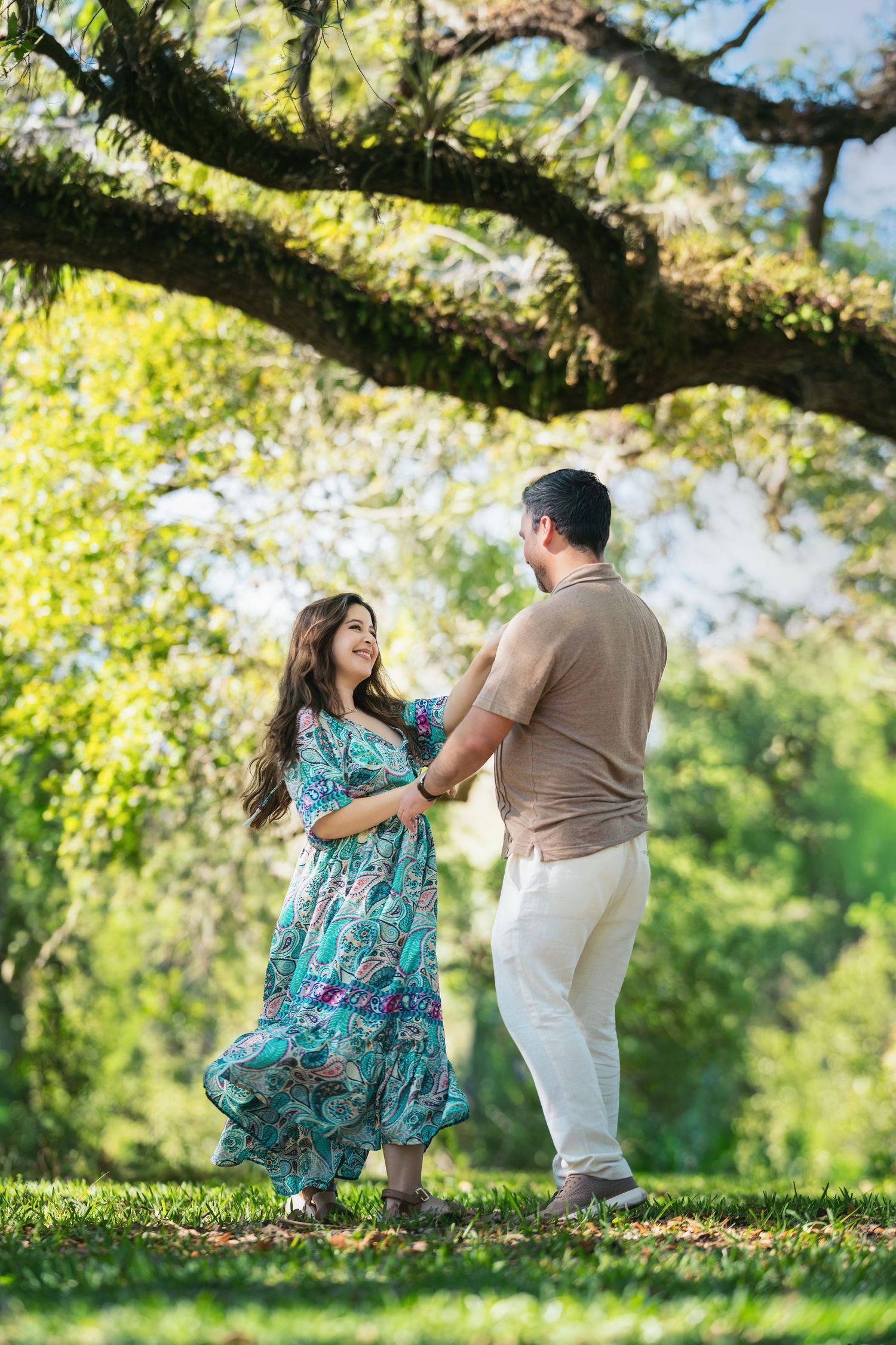 A couple dances joyfully under a moss-covered tree in a lush green park, woman wearing a floral dress.