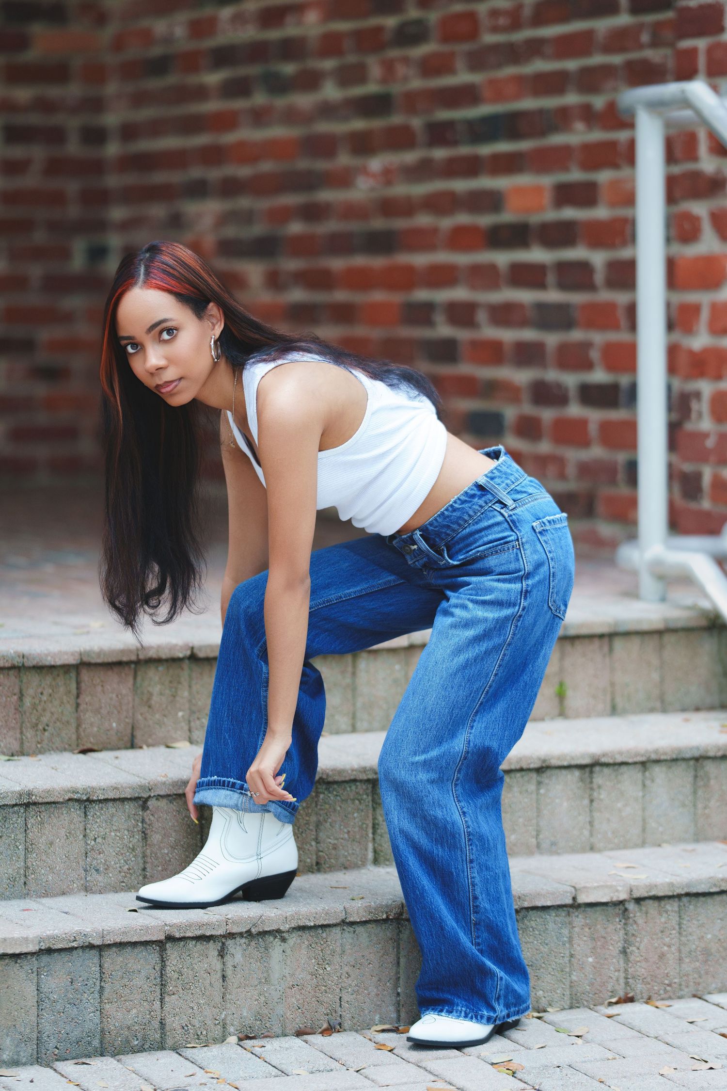 Young woman in white crop top, wide-leg jeans, and white cowboy boots posing on stone steps near brick wall.