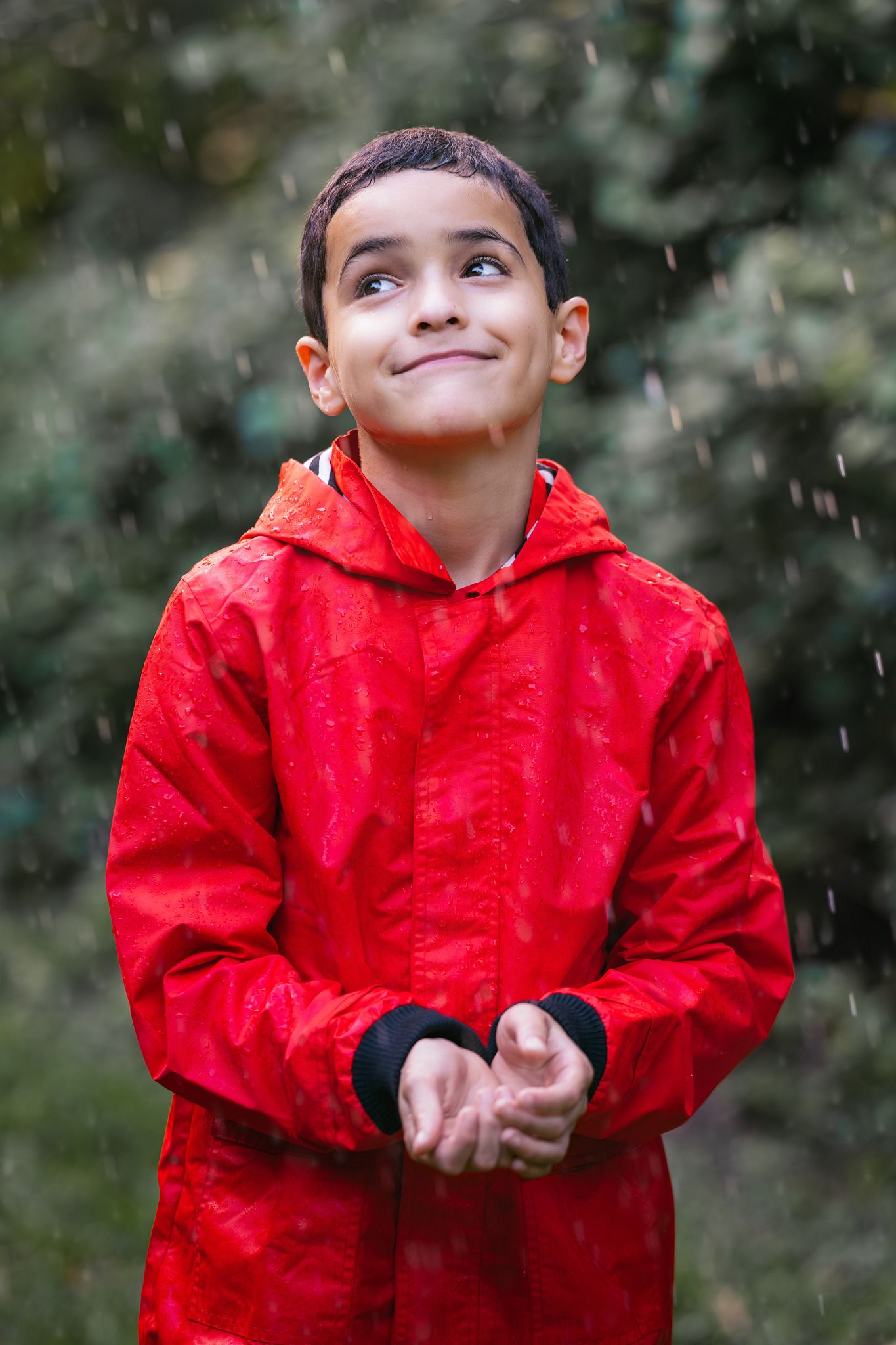 Young boy in red raincoat smiling and looking up at falling rain outdoors.
