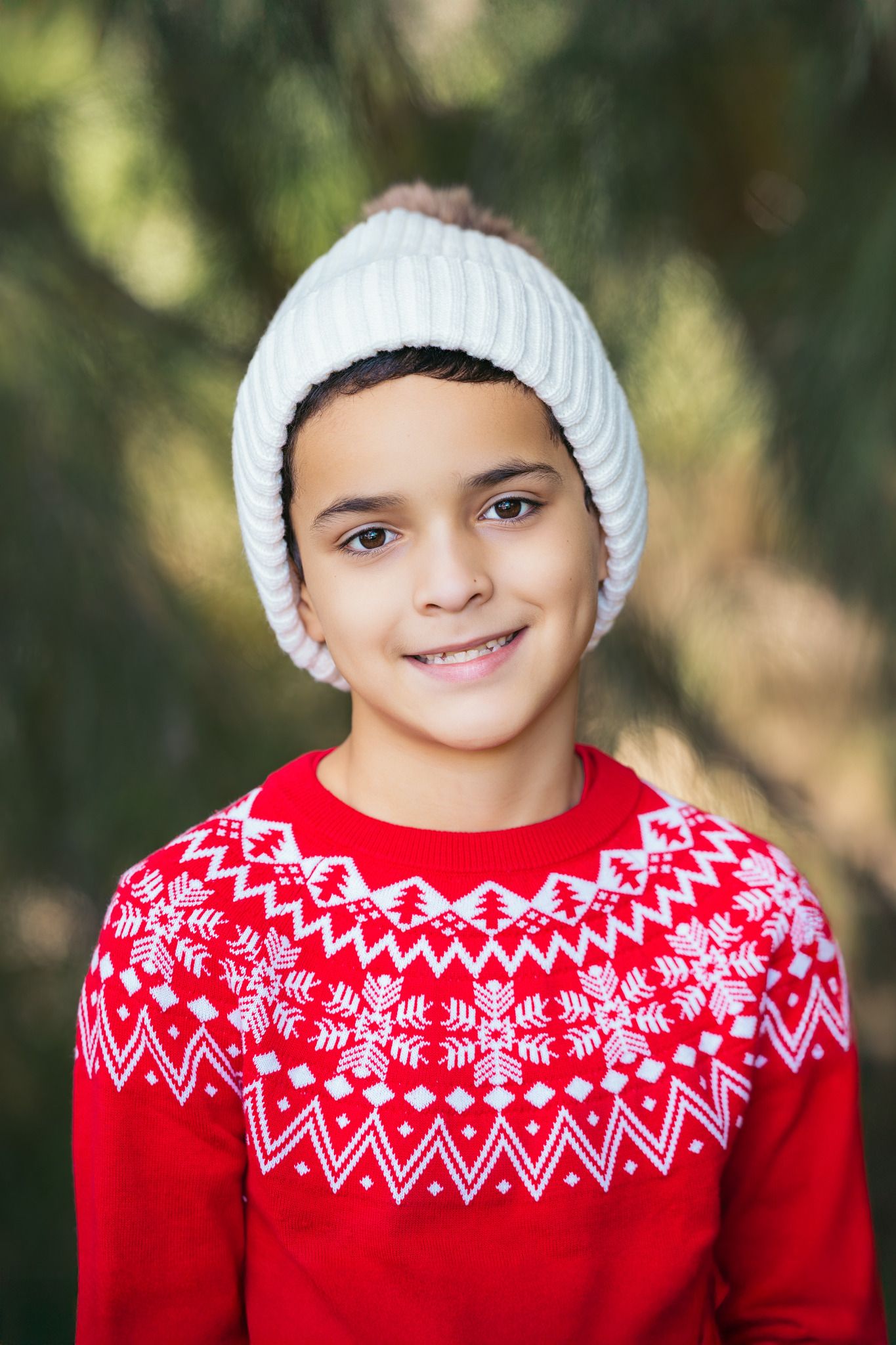 Smiling child wearing a white knit beanie and red Nordic Christmas sweater outdoors near green pine trees.