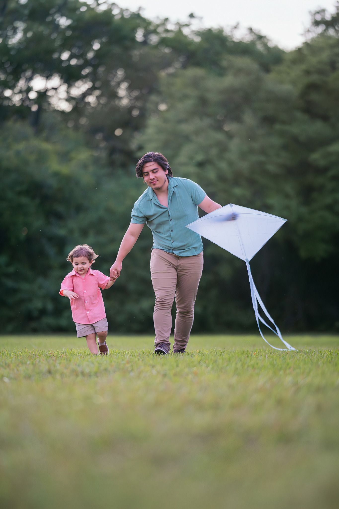 A mother and young child run together in a green park, flying a white kite on a warm summer day.