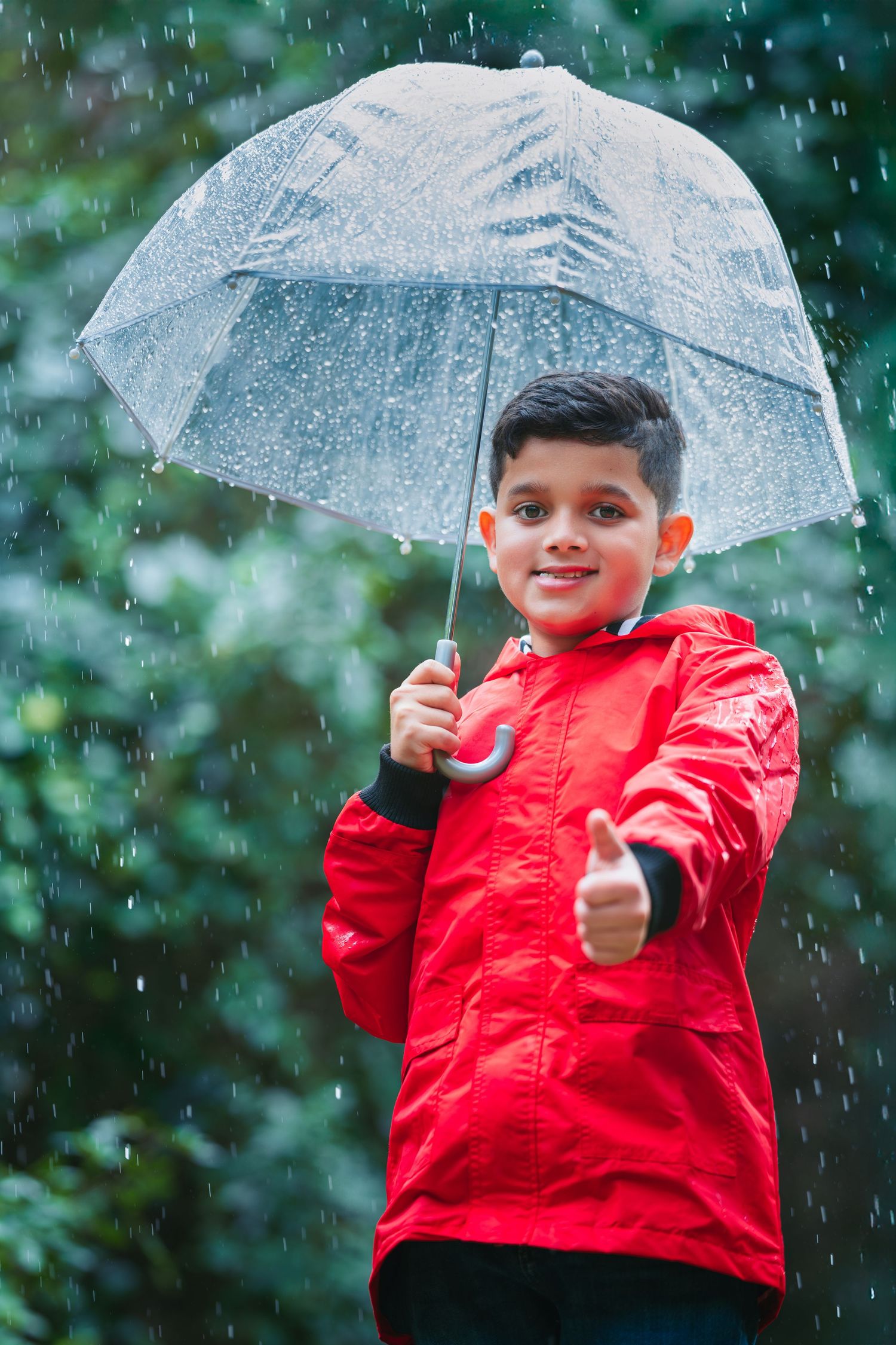Smiling boy in red raincoat holds clear umbrella giving thumbs up in the rain with green trees behind.