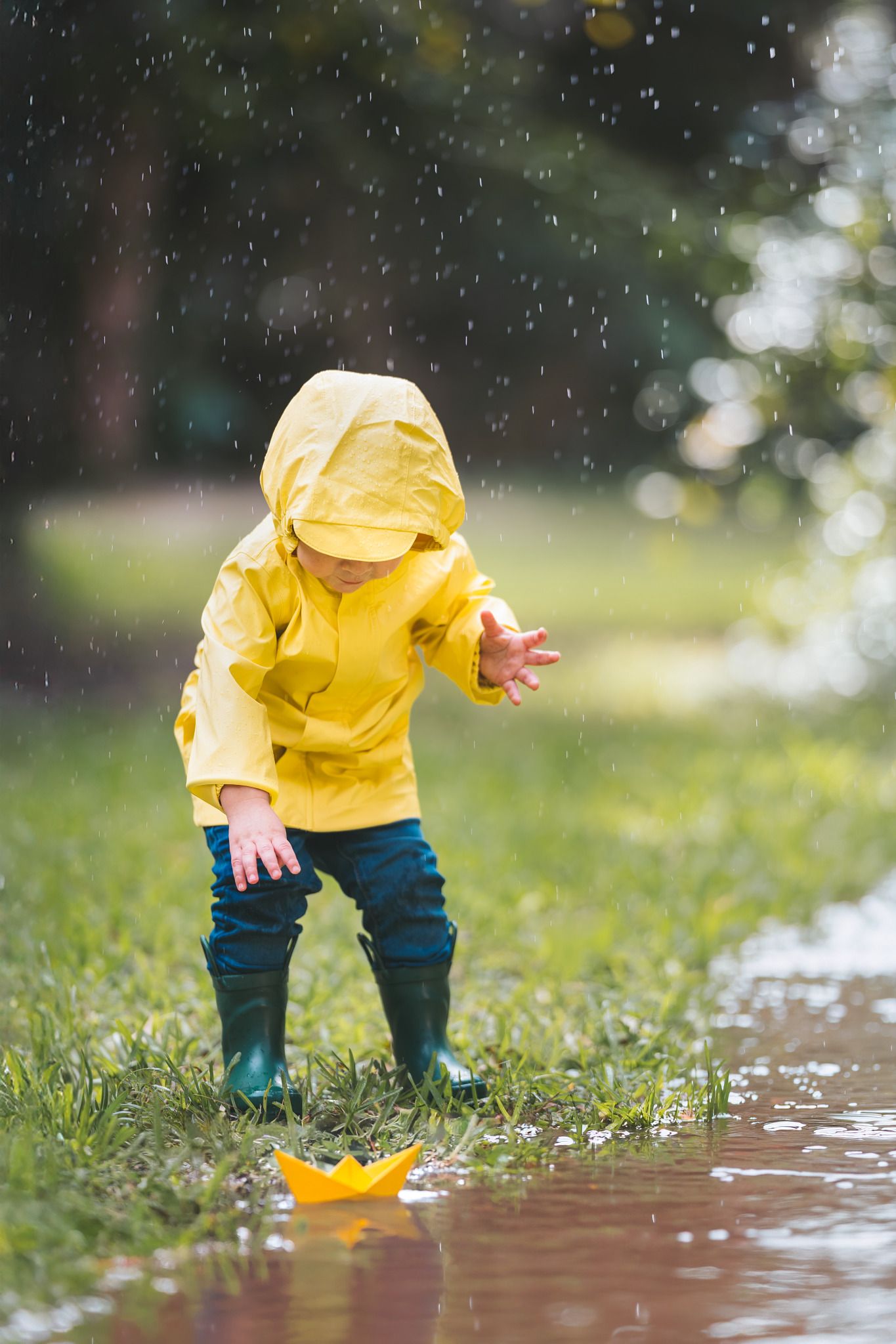 Toddler in yellow raincoat and boots playing in puddle with paper boat in the rain.