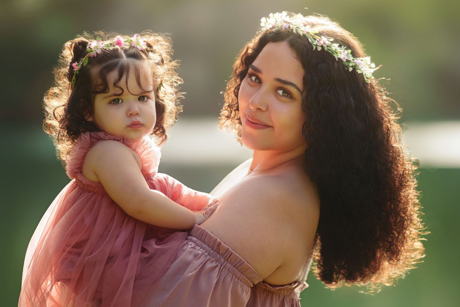 A woman with curly hair and floral crown holds a toddler girl in pink dresses outdoors in soft golden light.