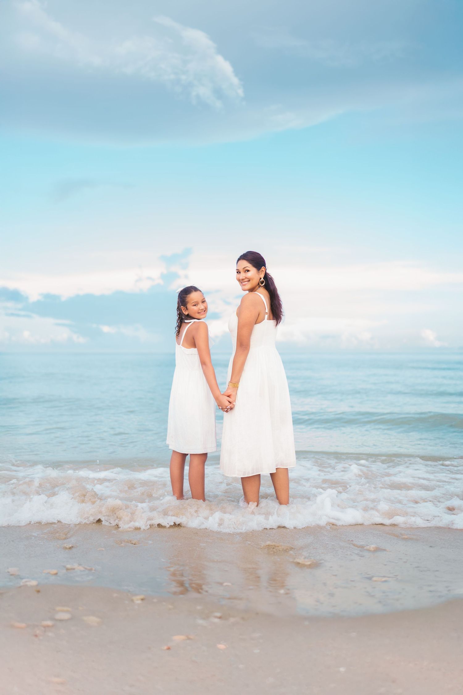 Mother and daughter in white dresses holding hands on a sandy beach with ocean waves and blue sky.