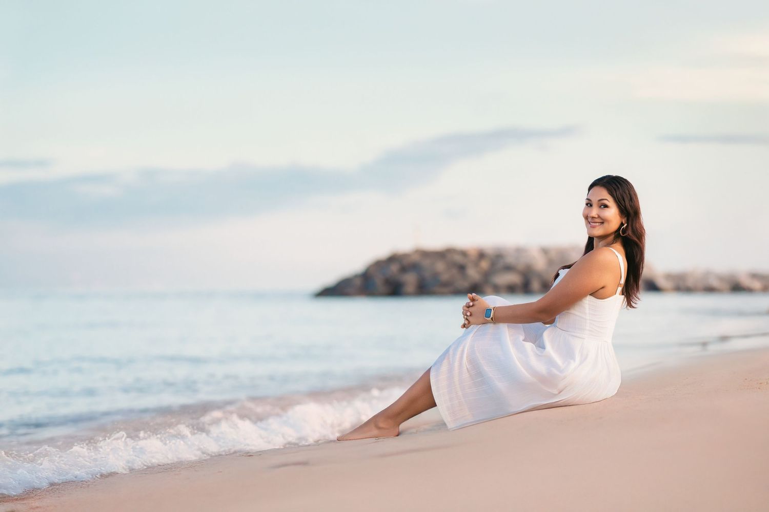 Woman in white dress sitting on the beach with ocean backdrop.