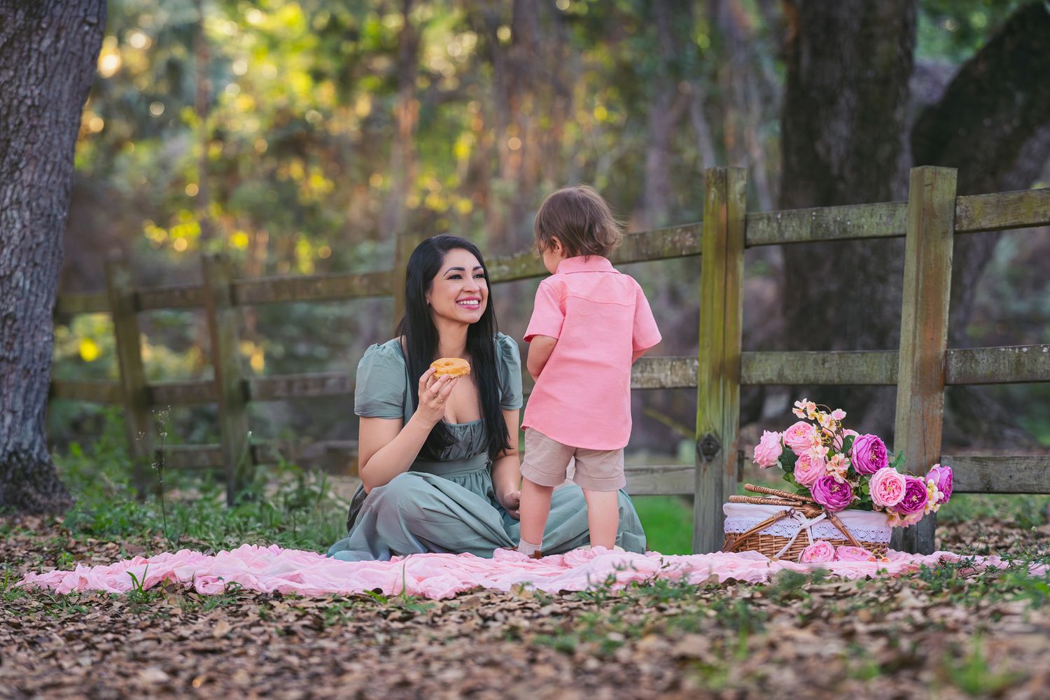 Woman and toddler enjoying a picnic on pink blanket outdoors near wooden fence surrounded by pink flowers.