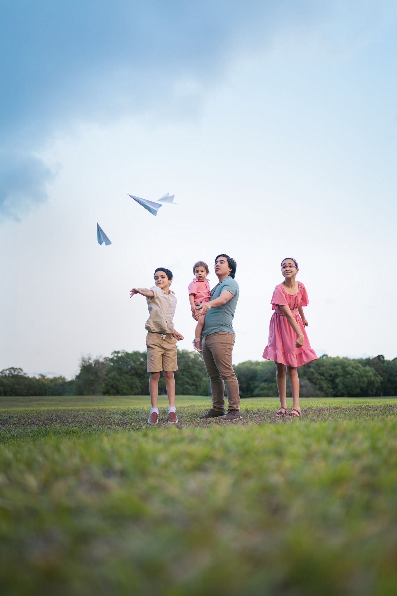 Family of four playing with colorful paper airplanes in a green park under a cloudy sky.