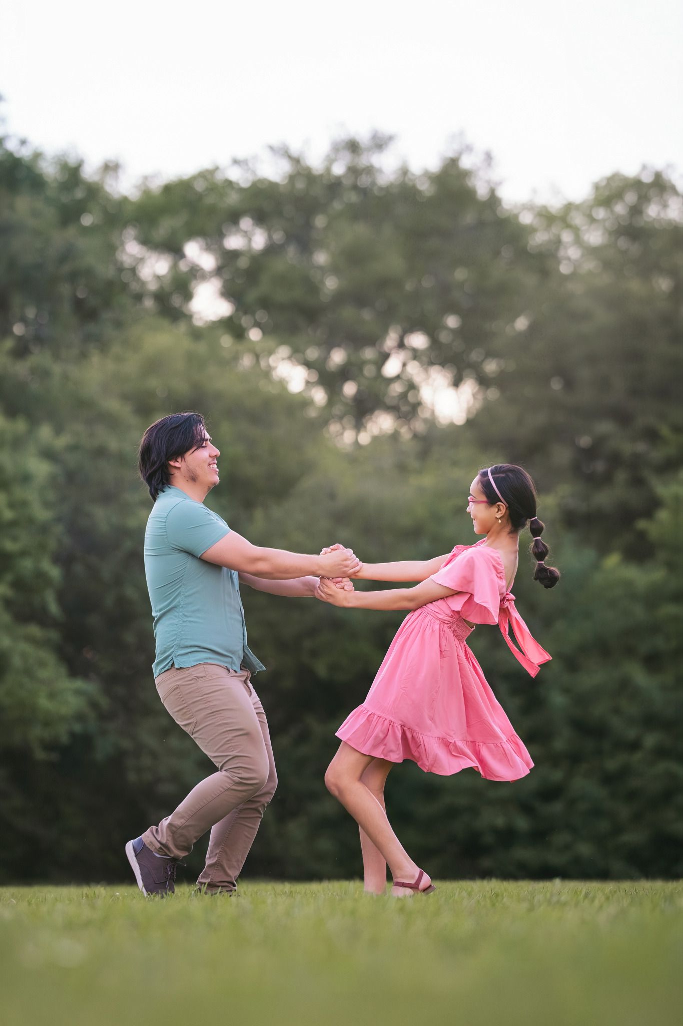 A couple joyfully spin together holding hands on a green lawn with lush trees behind them.