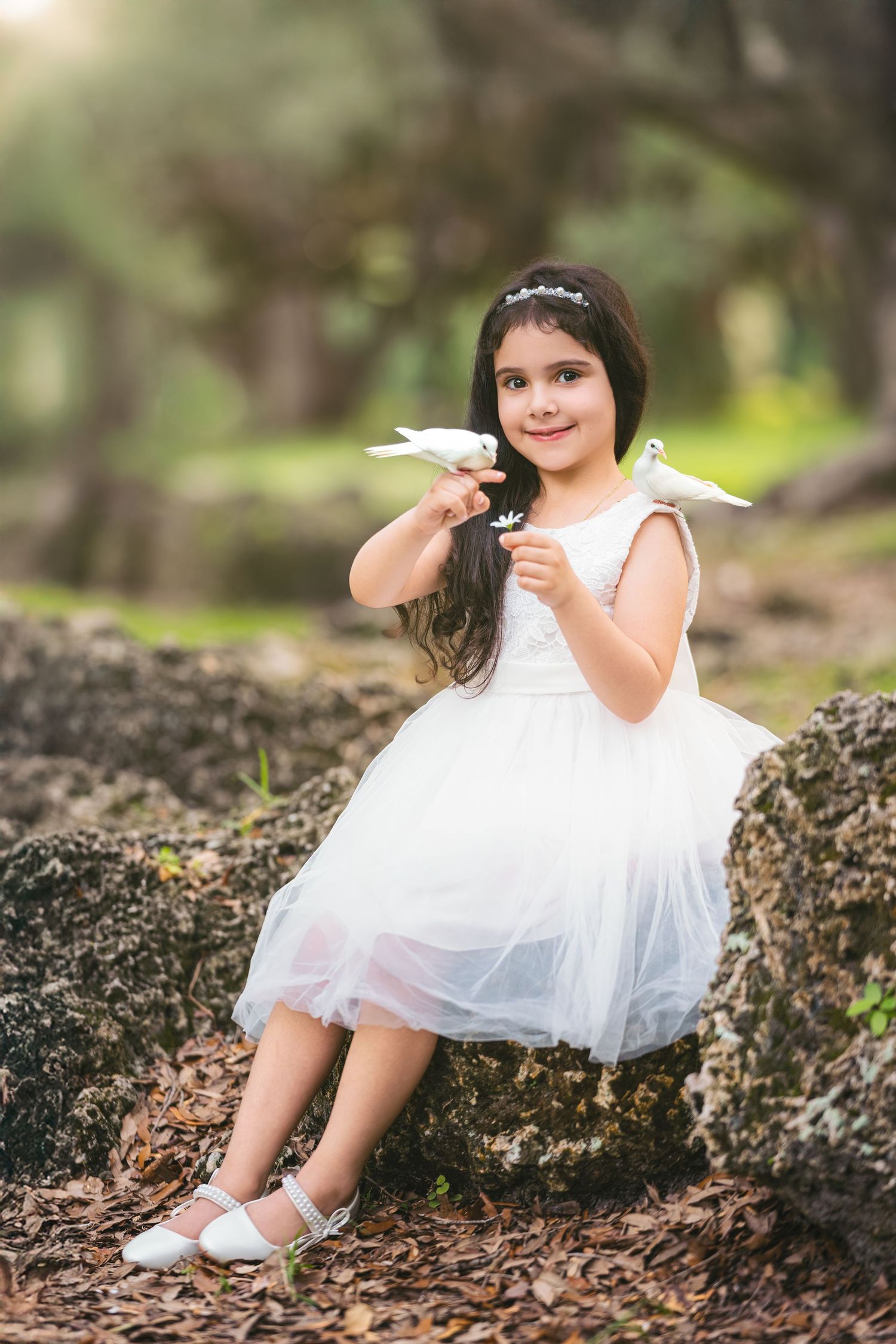 Young girl in white tulle dress sits on rocks holding white doves in a sunlit forest setting.
