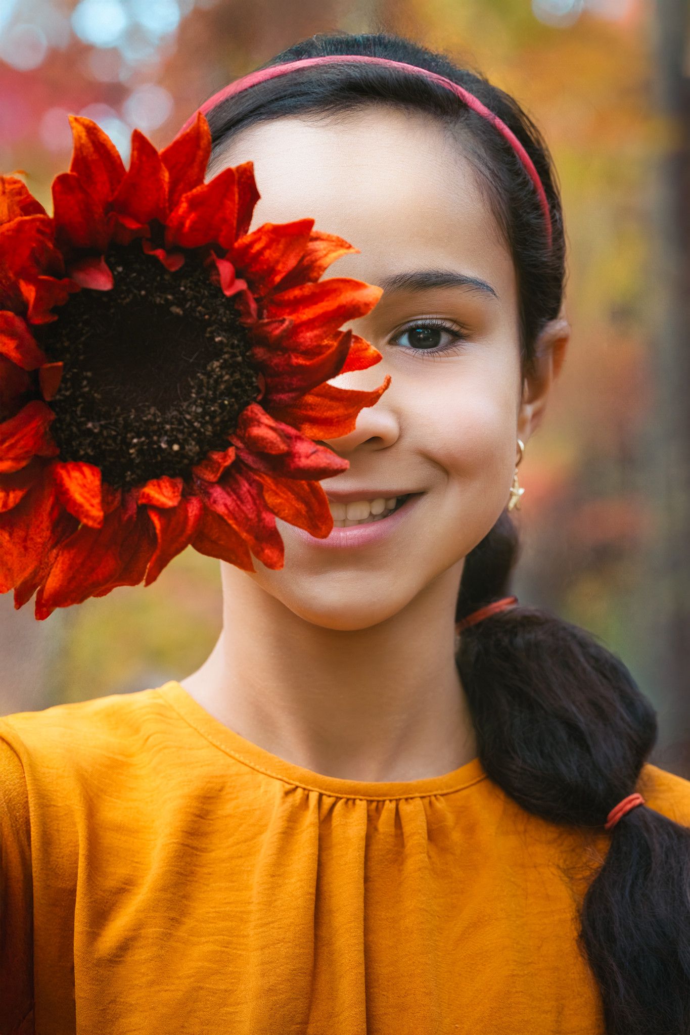 Smiling girl in yellow top holds red sunflower over one eye in autumn park setting.