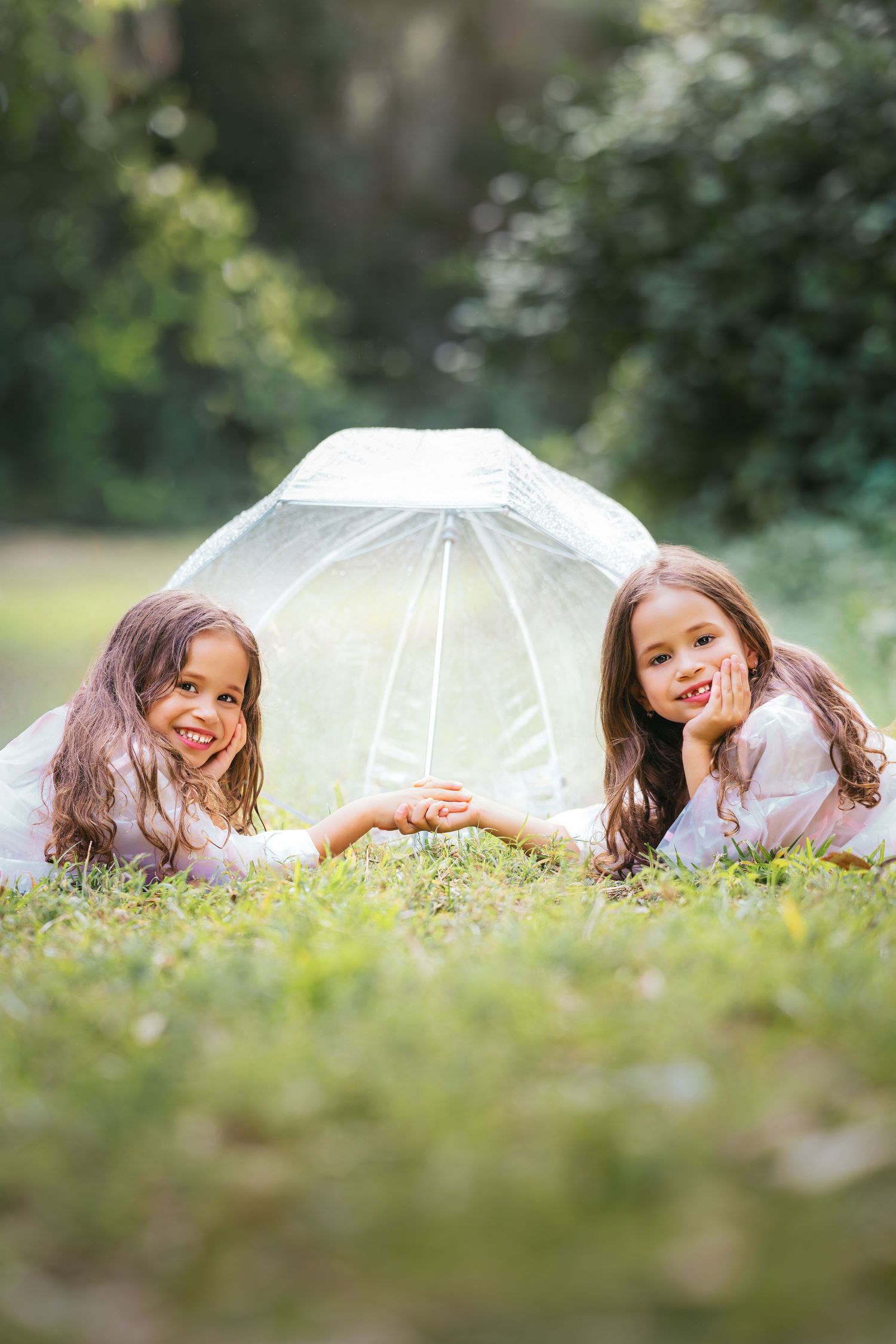 Two young girls lying on grass under a white transparent umbrella, smiling and holding hands in a lush green park.