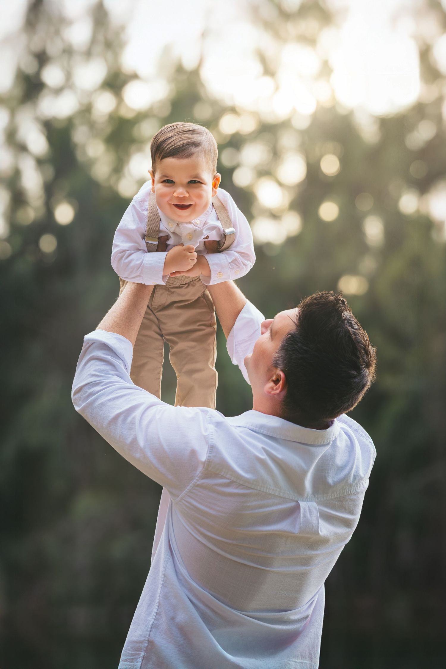 Father lifting smiling baby boy in white shirt outdoors with green trees in sunlit background.