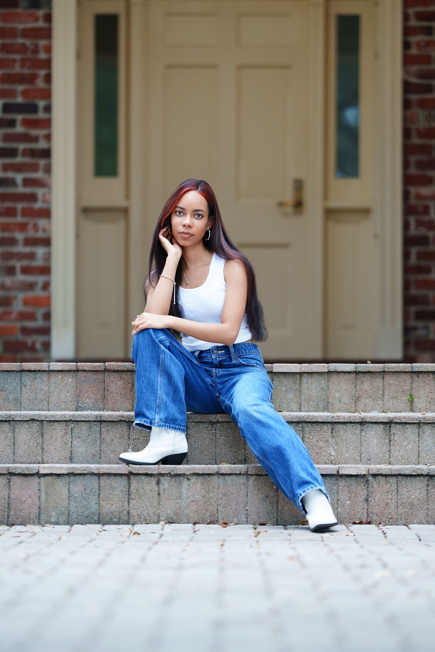 Young woman in white tank top, blue jeans, and white boots sitting on brick steps in front of a wooden door.