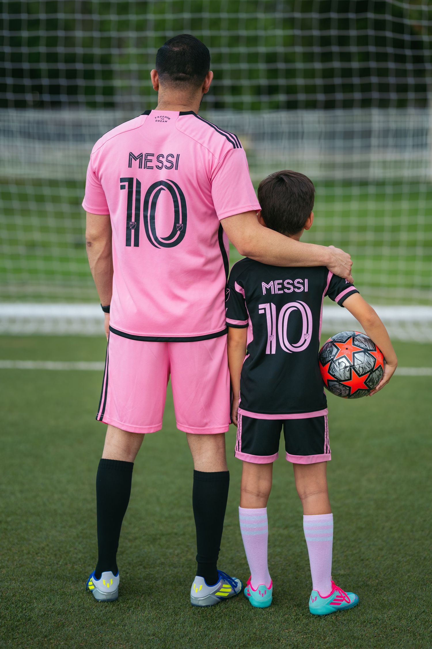 Two young soccer players wearing pink jerseys stand together on a field with a soccer ball.