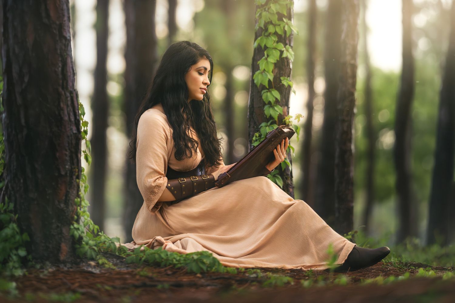 Young woman in beige dress reading a book while sitting against a tree in a misty forest.