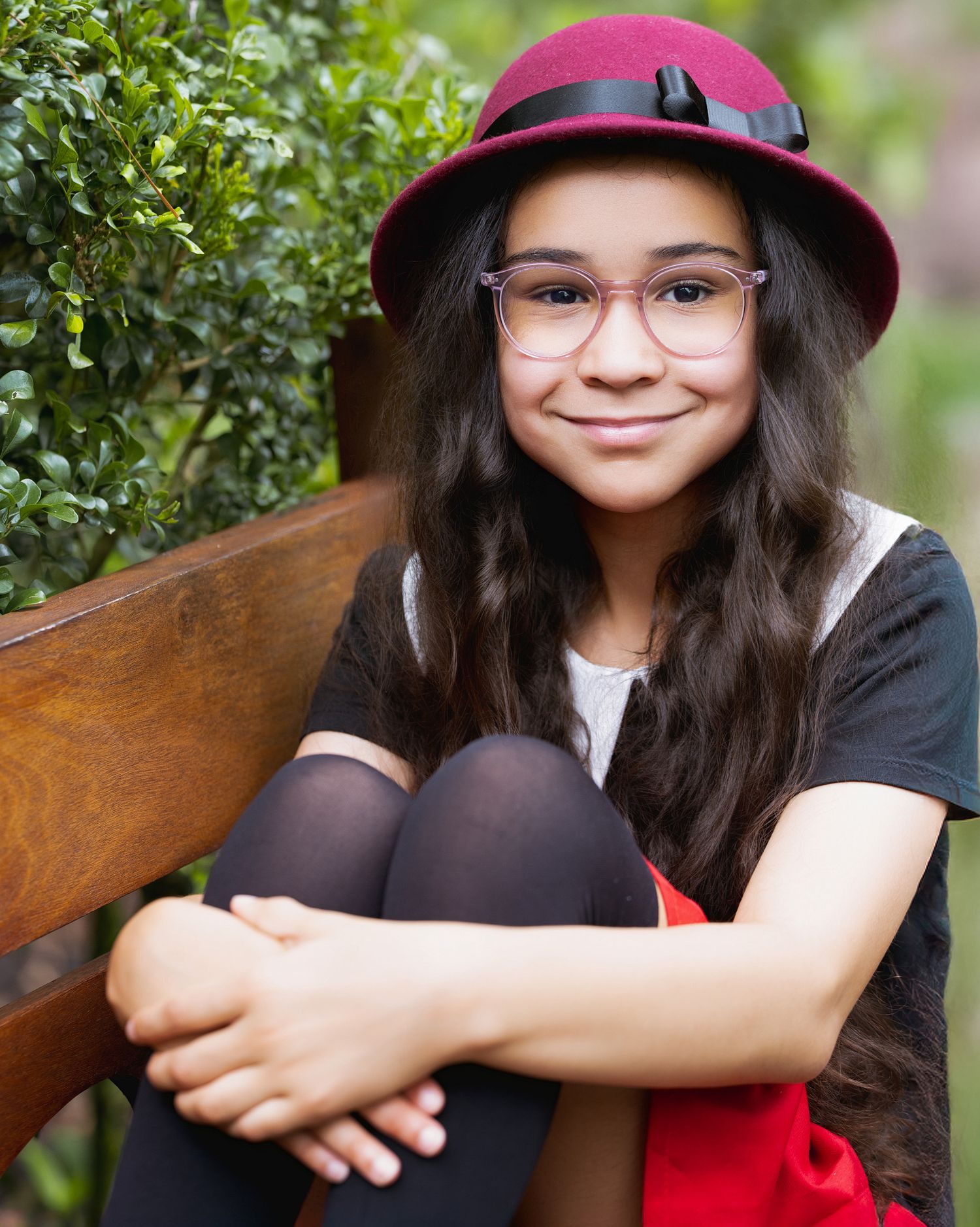 Someone wearing glasses and a pink helmet sits on a wooden bench surrounded by greenery.