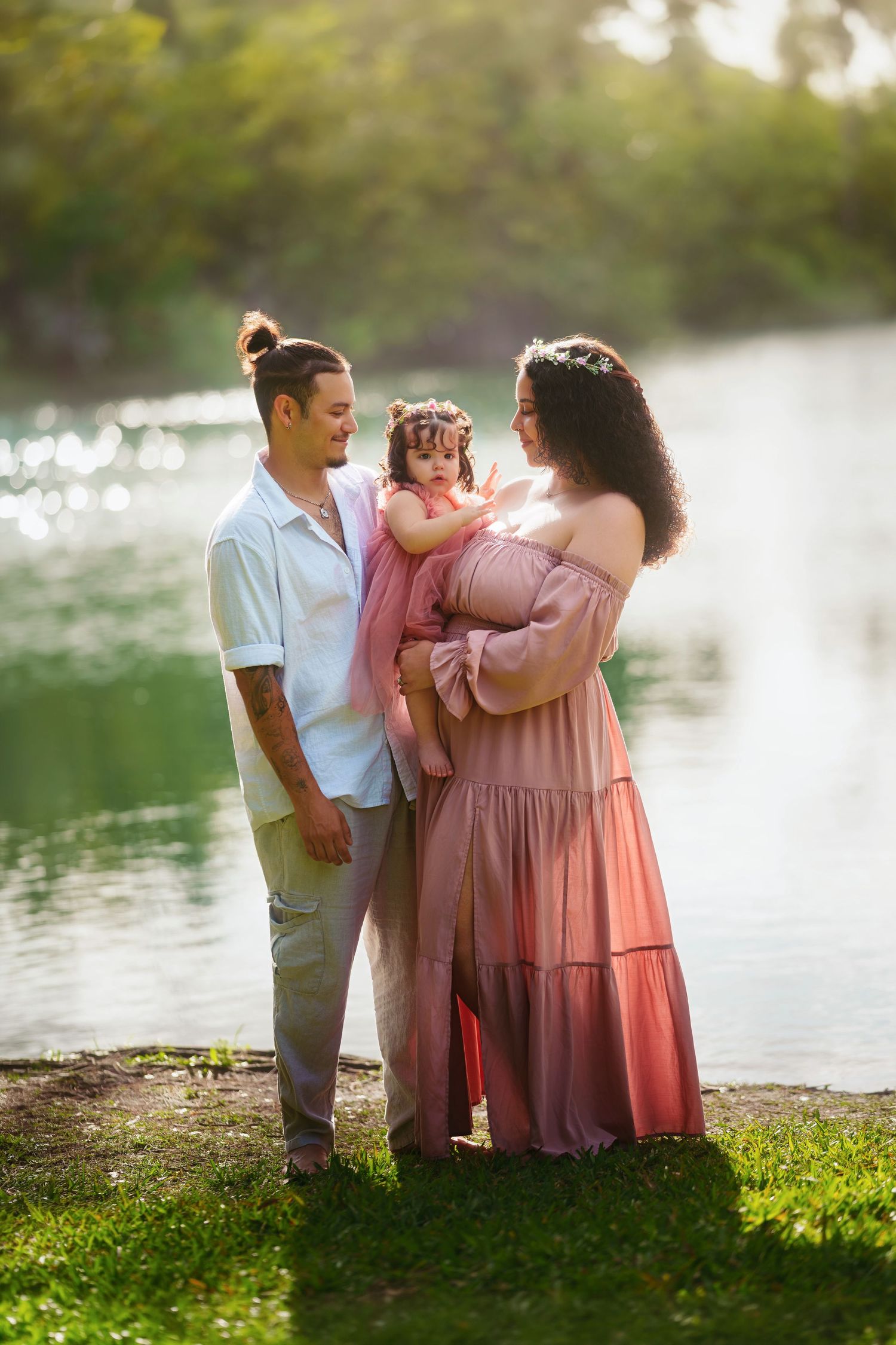 A young family with a toddler poses by a sunlit lake, mom in a pink maxi dress, dad in casual attire.