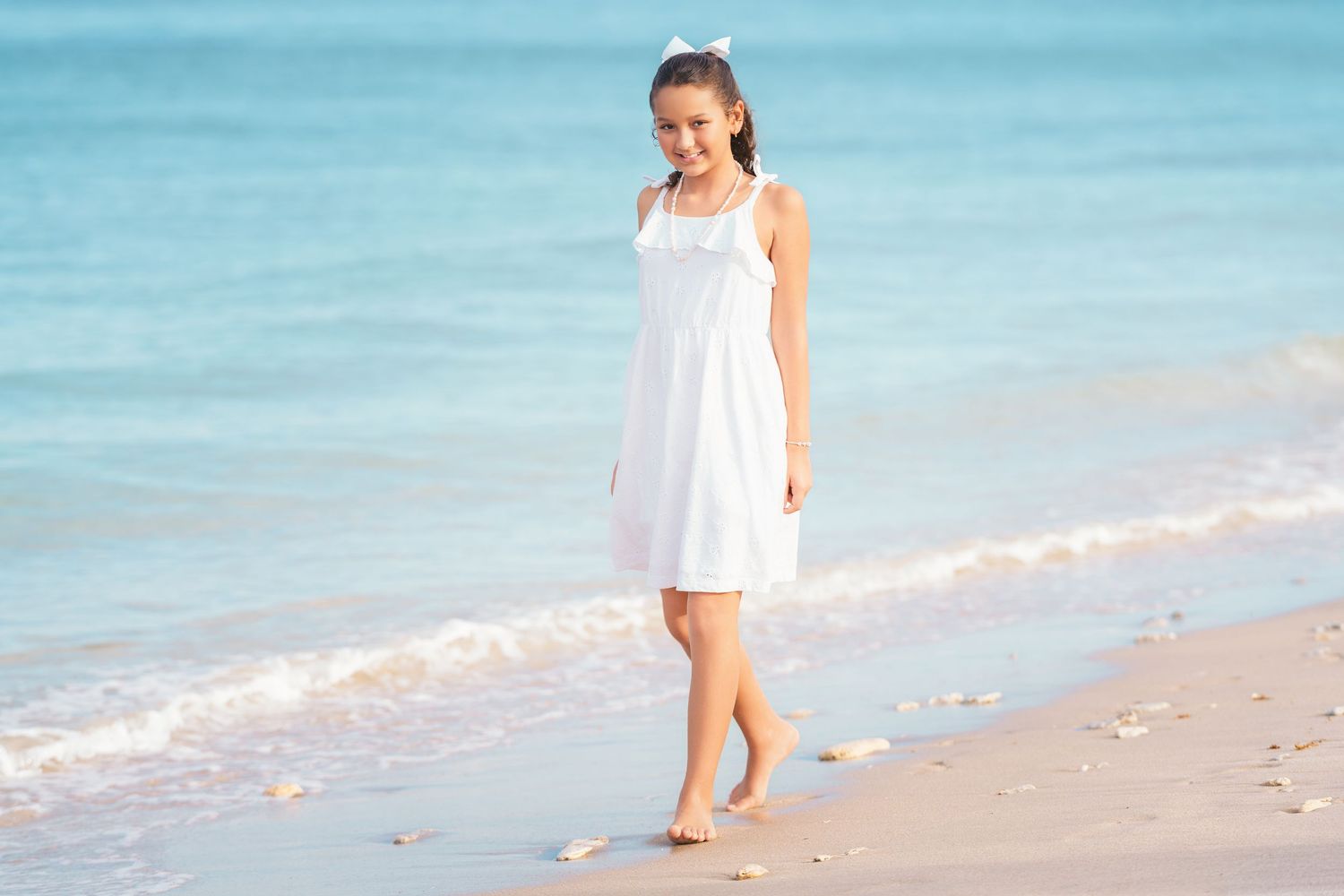 Young girl in white dress walking barefoot on a sunny beach with gentle ocean waves.