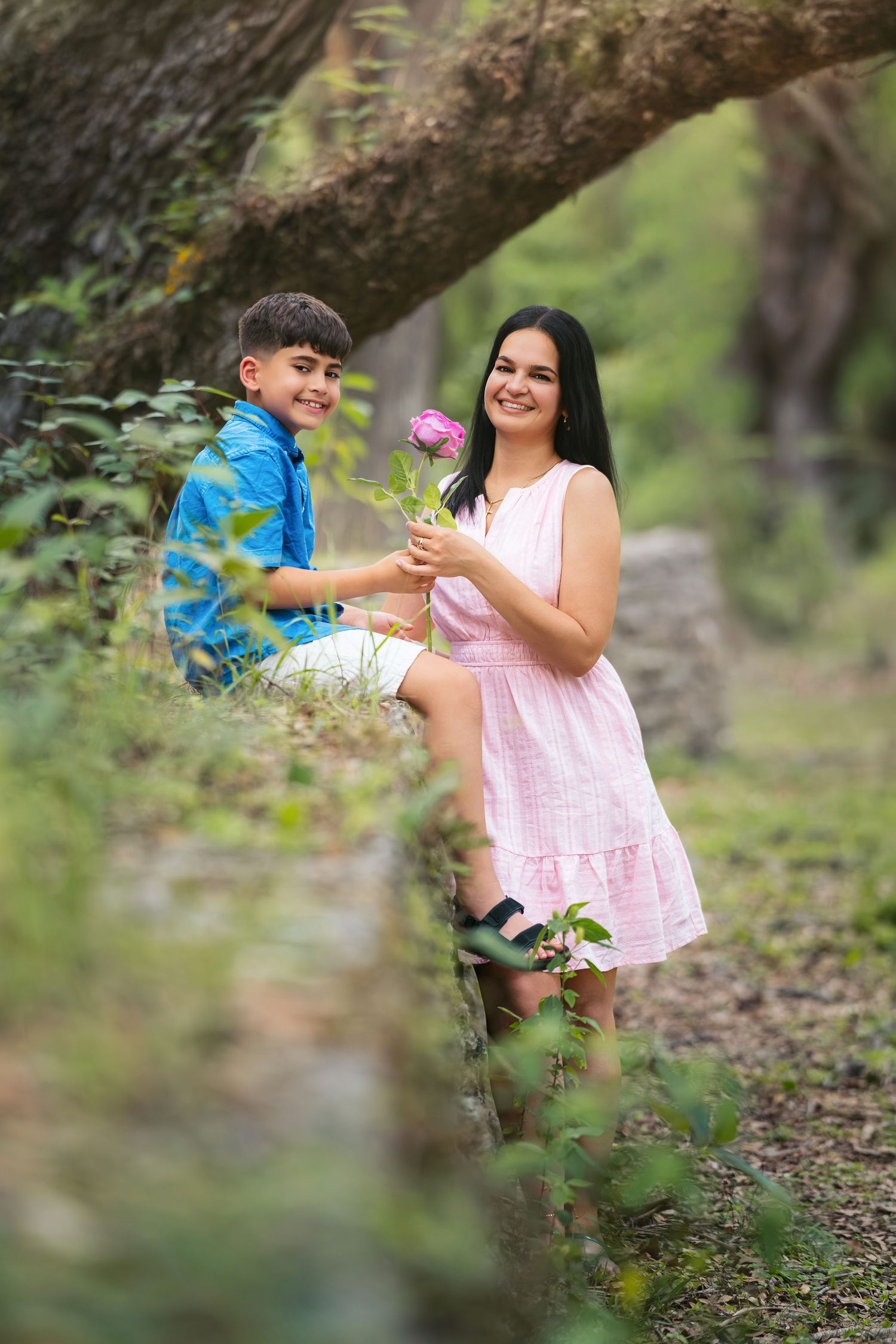 A boy in a blue shirt offers a pink flower to a smiling girl in a pink dress outdoors.