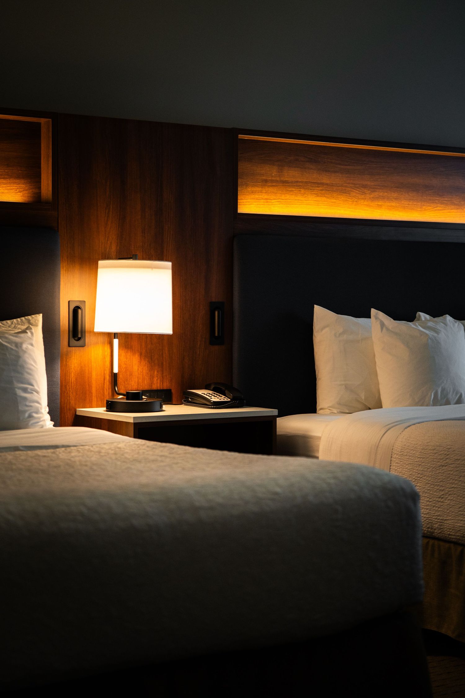 Modern hotel room interior with two beds illuminated by warm ambient lighting and bedside lamp on wooden paneled walls.
