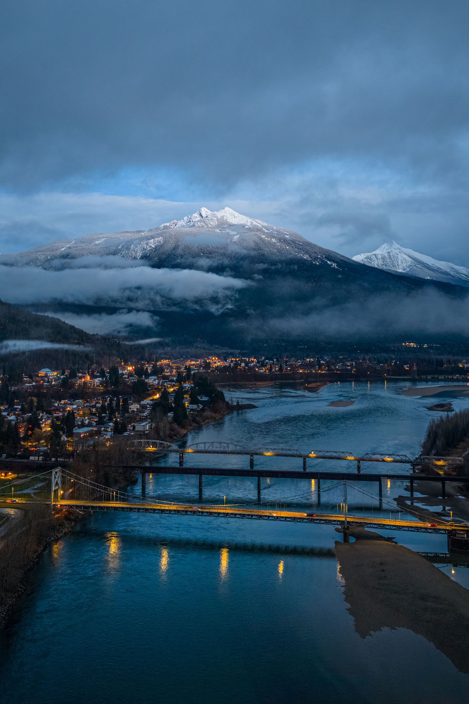 Panoramic dusk view of a mountain town with snow-capped peaks, illuminated bridges and misty waters in the foreground.