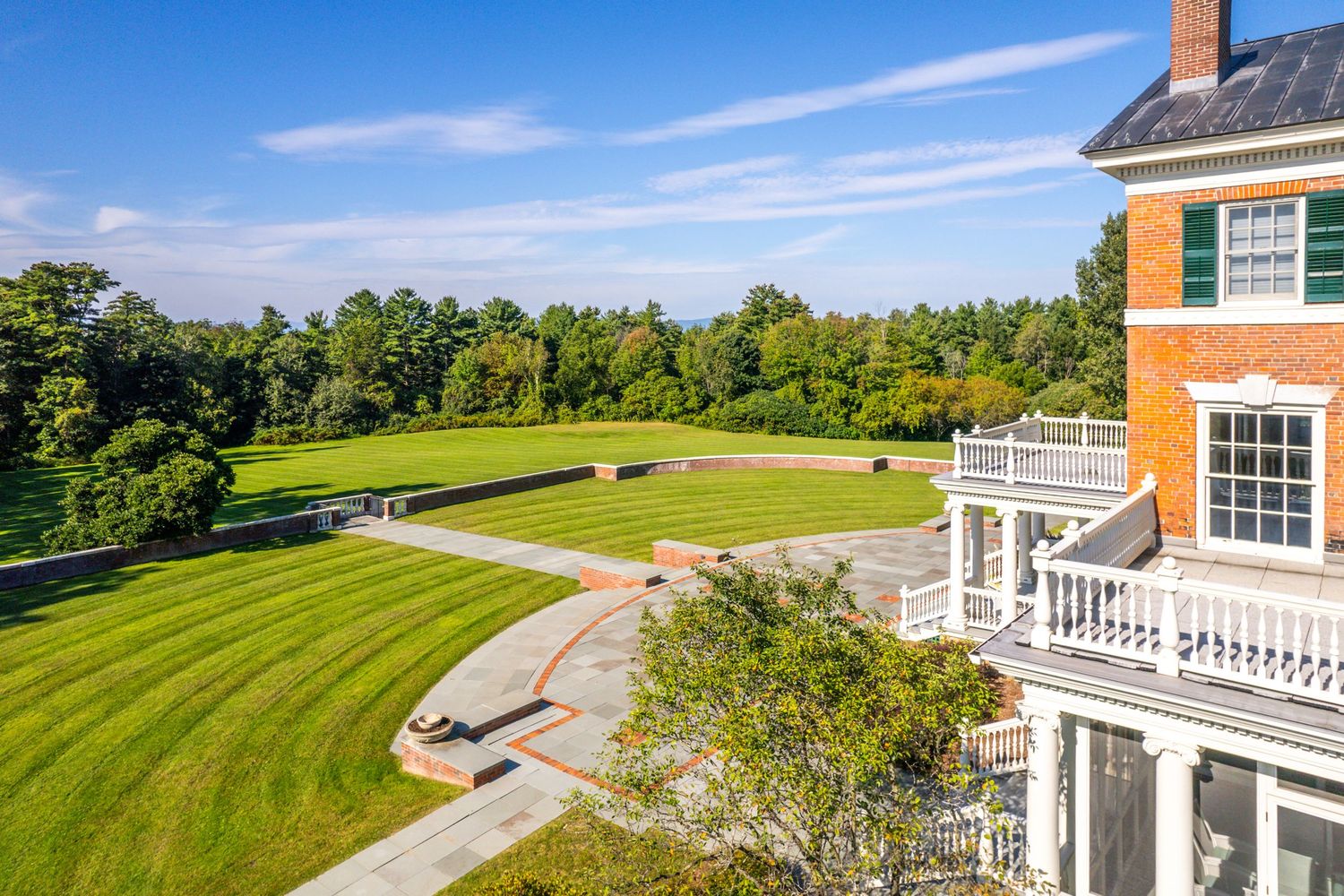 Drone shot of an elegant brick mansion with white trim and curved driveway nestled among green lawns.