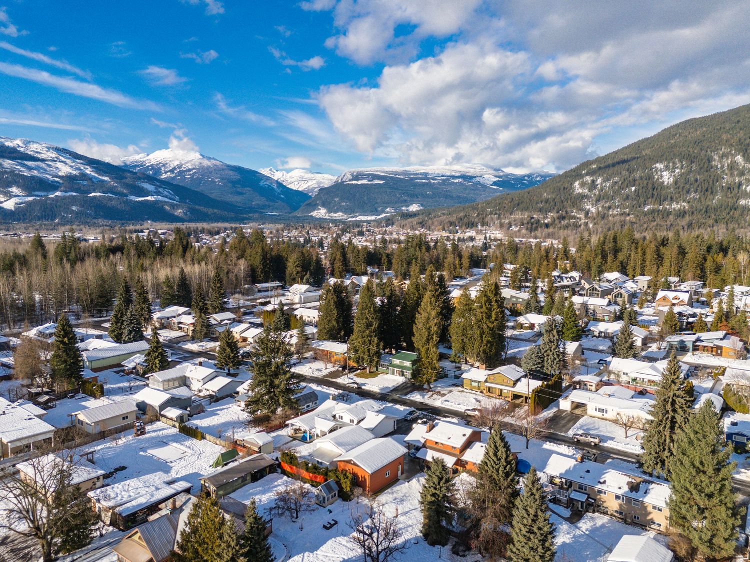 Aerial panoramic view of a snow-covered mountain town with chalets and evergreen trees against a bright blue winter sky.