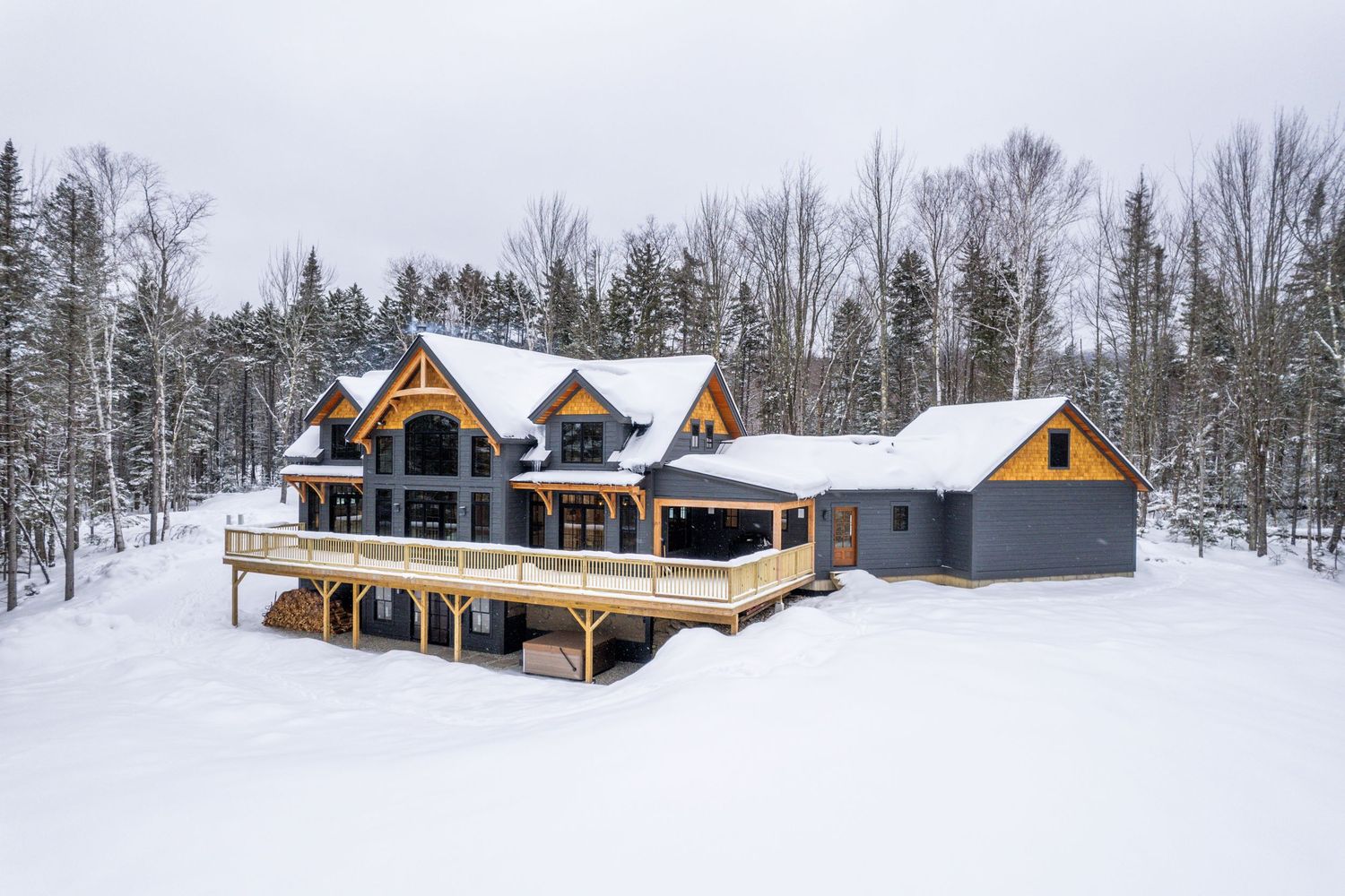 Snow-covered modern timber frame house with expansive deck and large windows in winter forest setting.