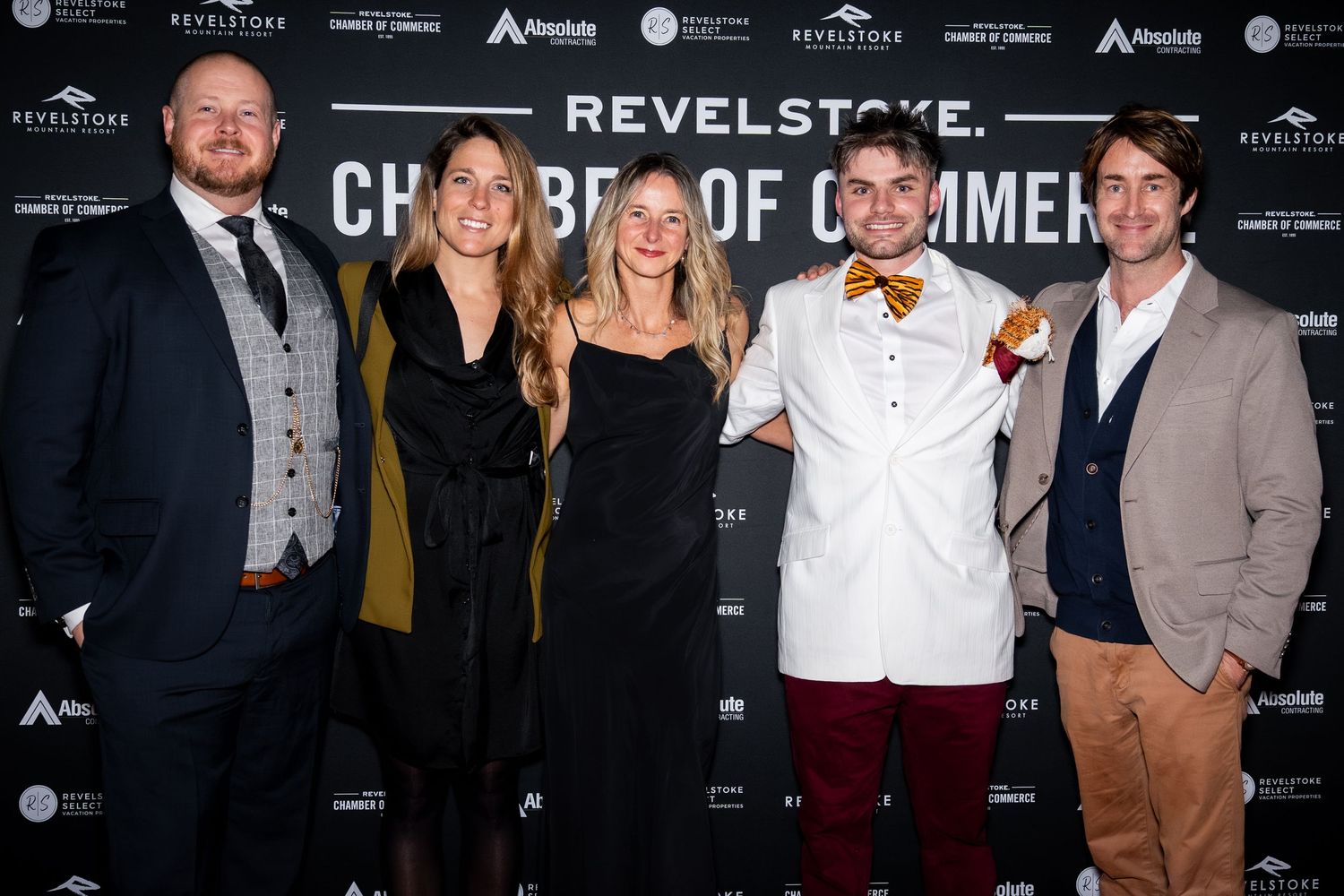 People in formal attire pose together at a red carpet event with promotional banners visible in the background.