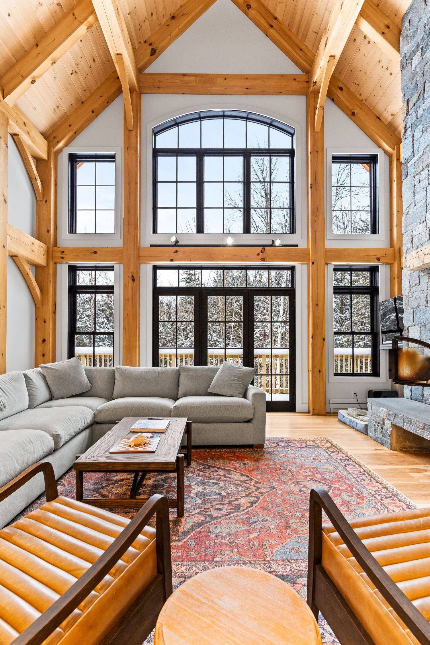 Double-height living room with exposed timber frame, stone fireplace, and wall of dramatic black windows.