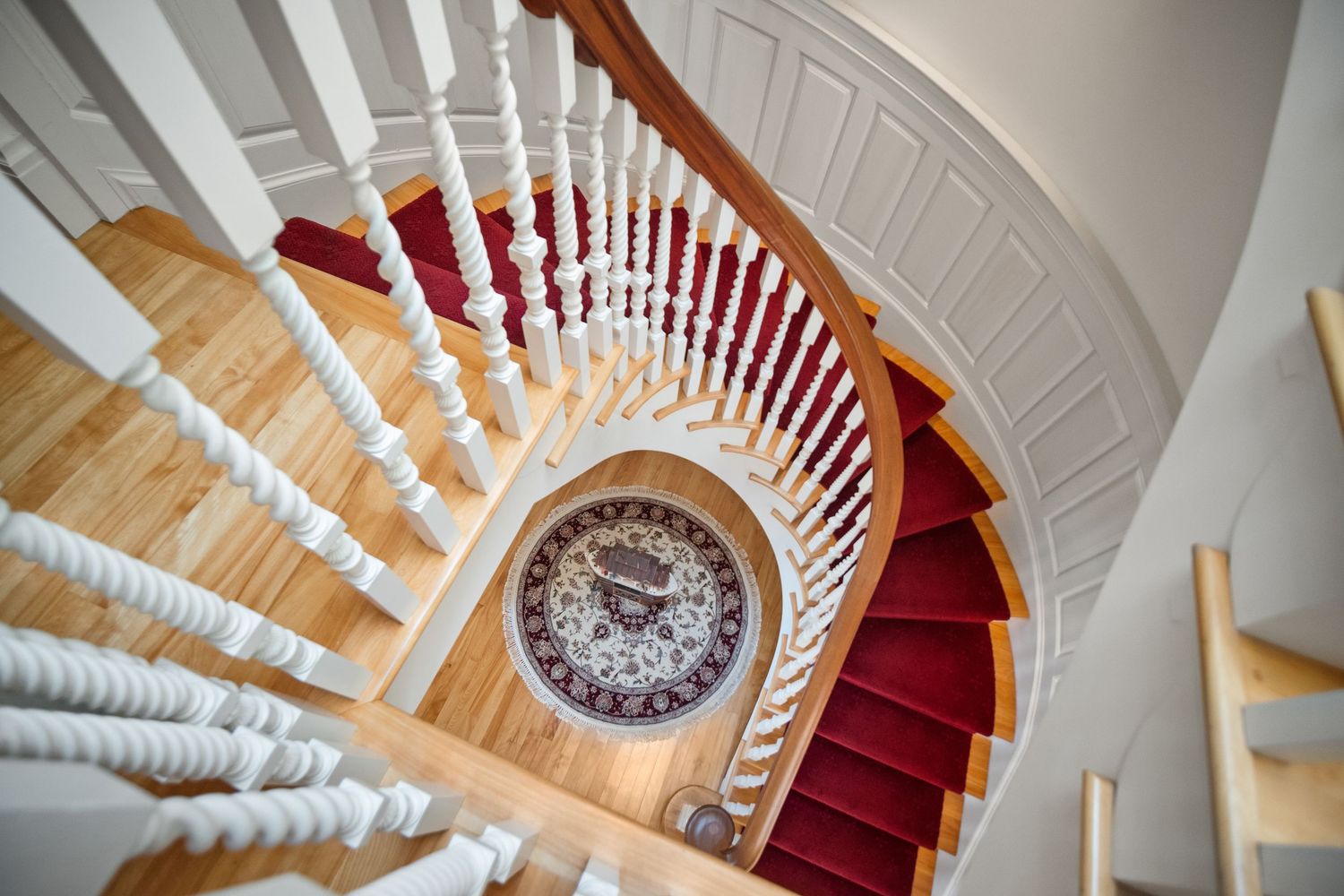 Elegant spiral staircase with white spindles, wooden treads, and red carpet runner viewed from above.