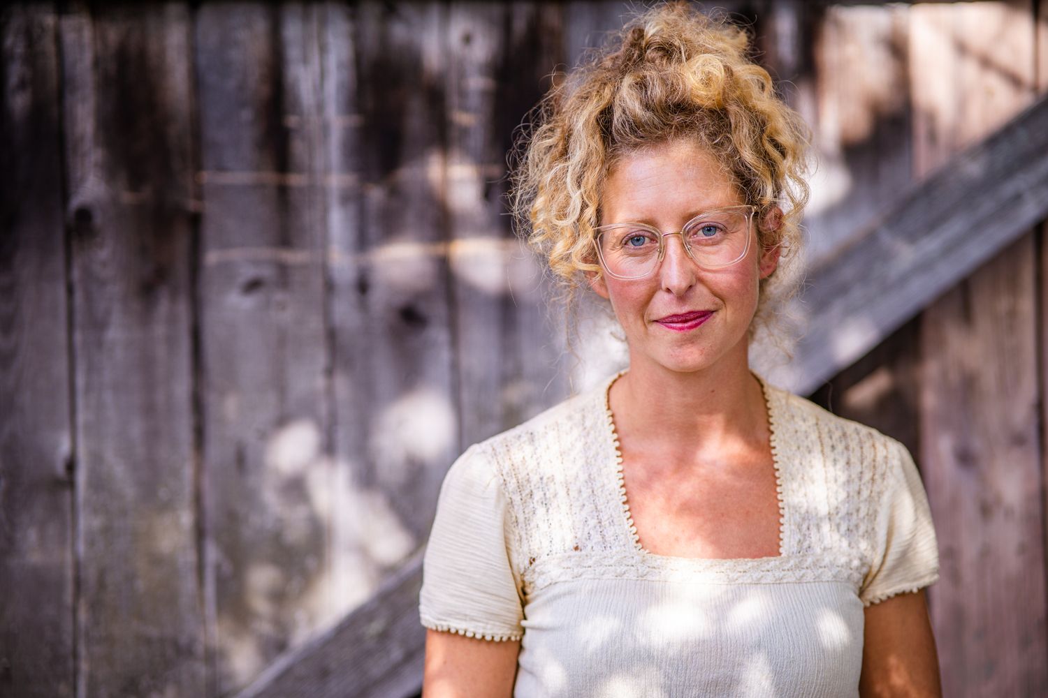 A person with curly blonde hair wearing a white lace top poses outdoors against a wooden fence background.