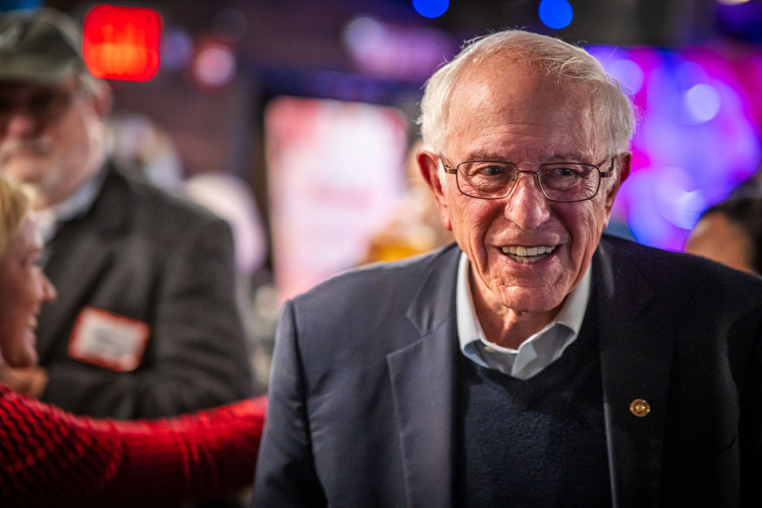 A person in a suit smiles during a colorful event with red and blue lighting in the background.