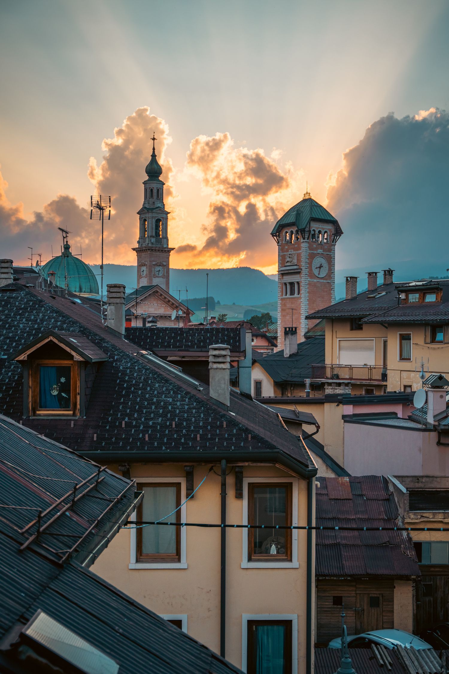 Dramatic sunset sky over European village rooftops with church spires and clock towers silhouetted against orange clouds.