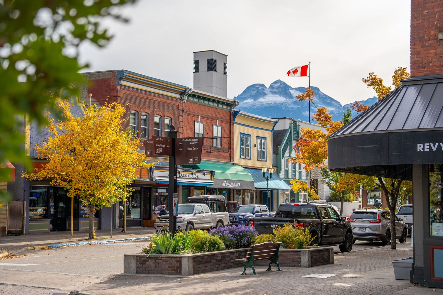 Panoramic view of a small mountain town's main street with fall foliage, brick buildings, and snow-capped peaks in distance.