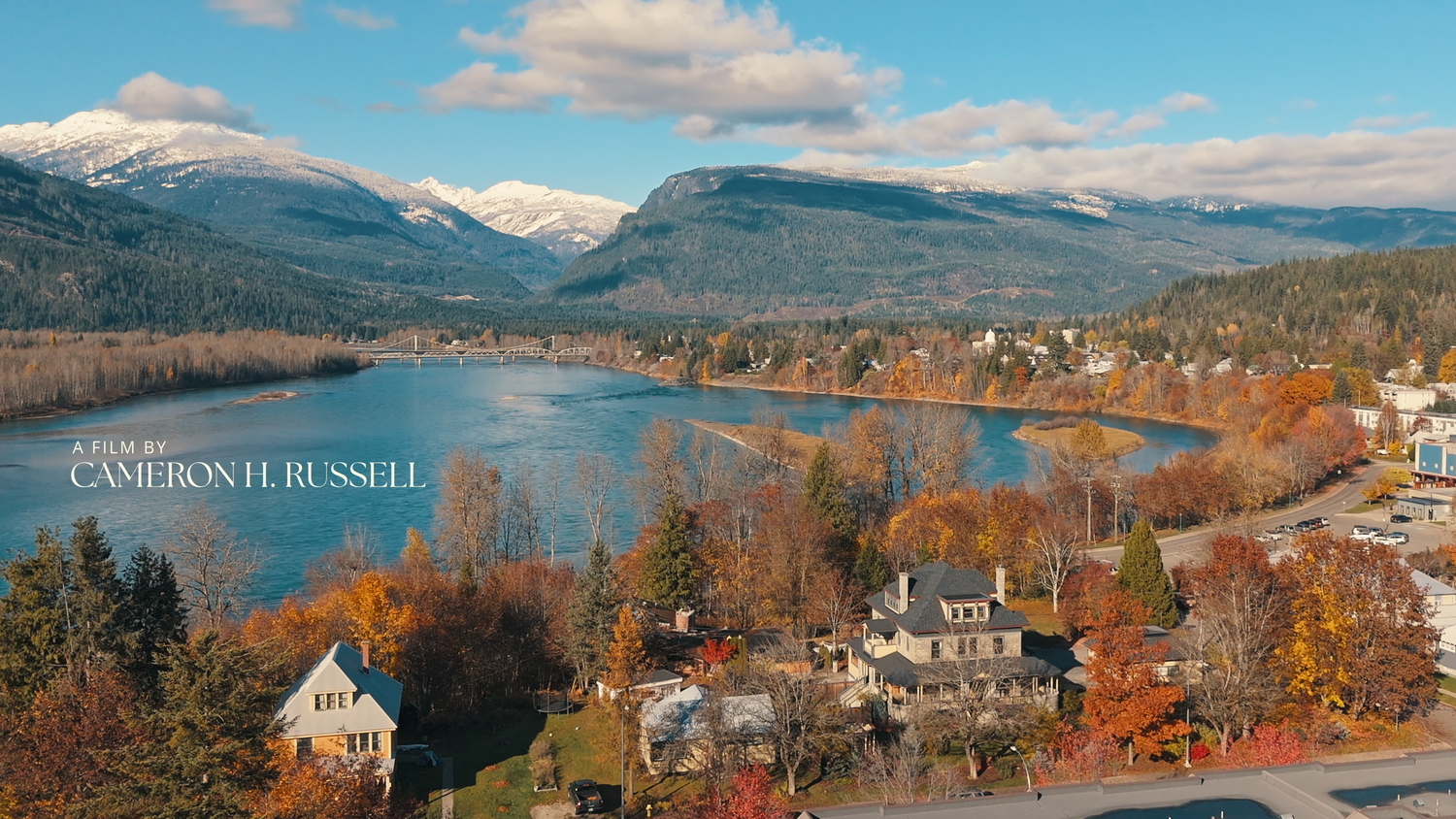A scenic panoramic view of a lakeside town nestled among autumn trees with snow-capped mountains in the distance.