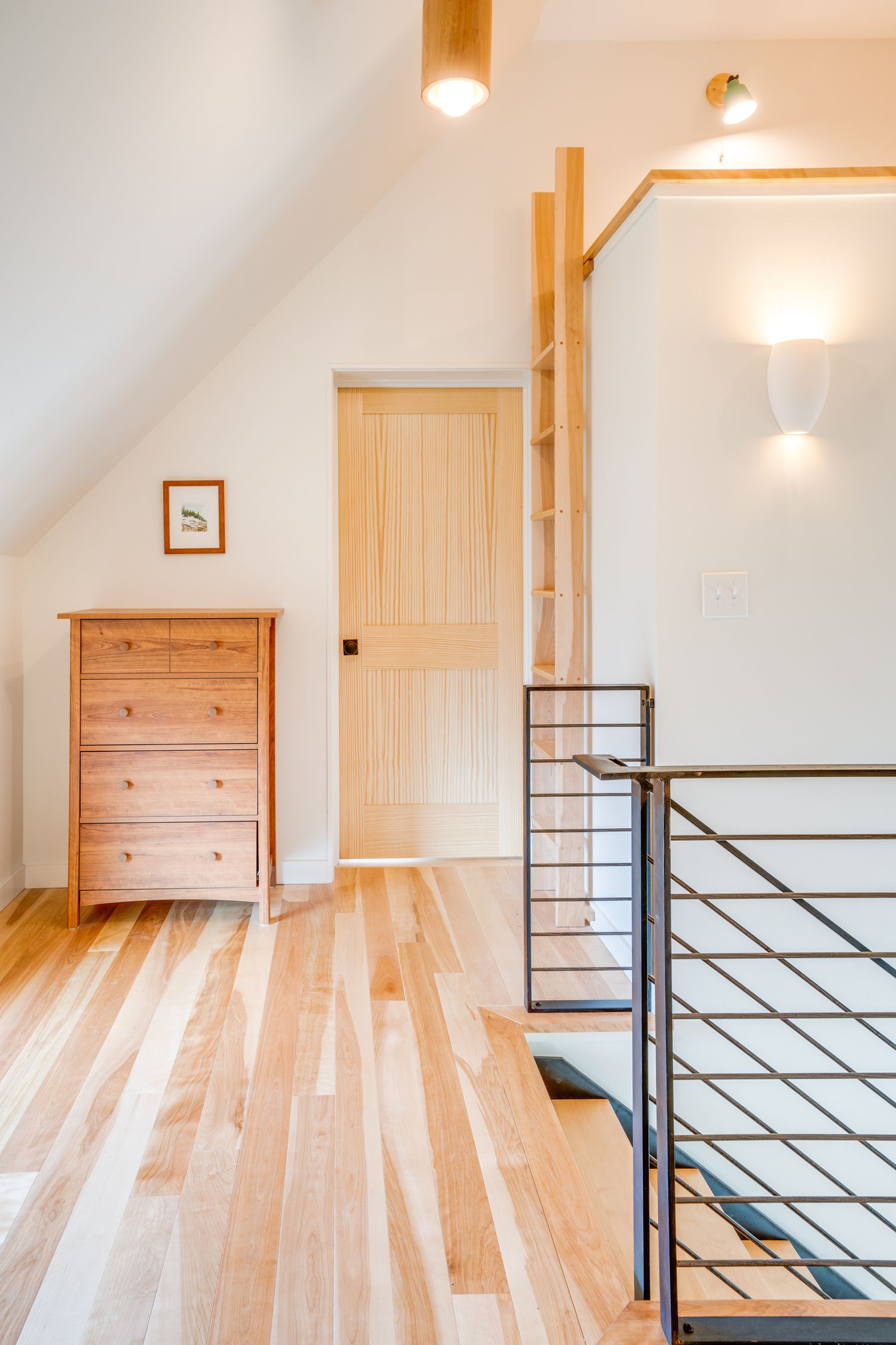 Bright upstairs hallway with wooden flooring, modern metal railing, and natural wood dresser.