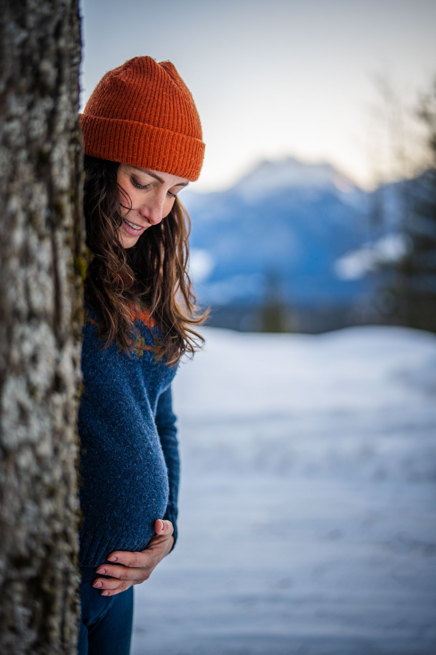 Person in orange beanie and blue sweater peeks from behind tree trunk in snowy winter mountain landscape.