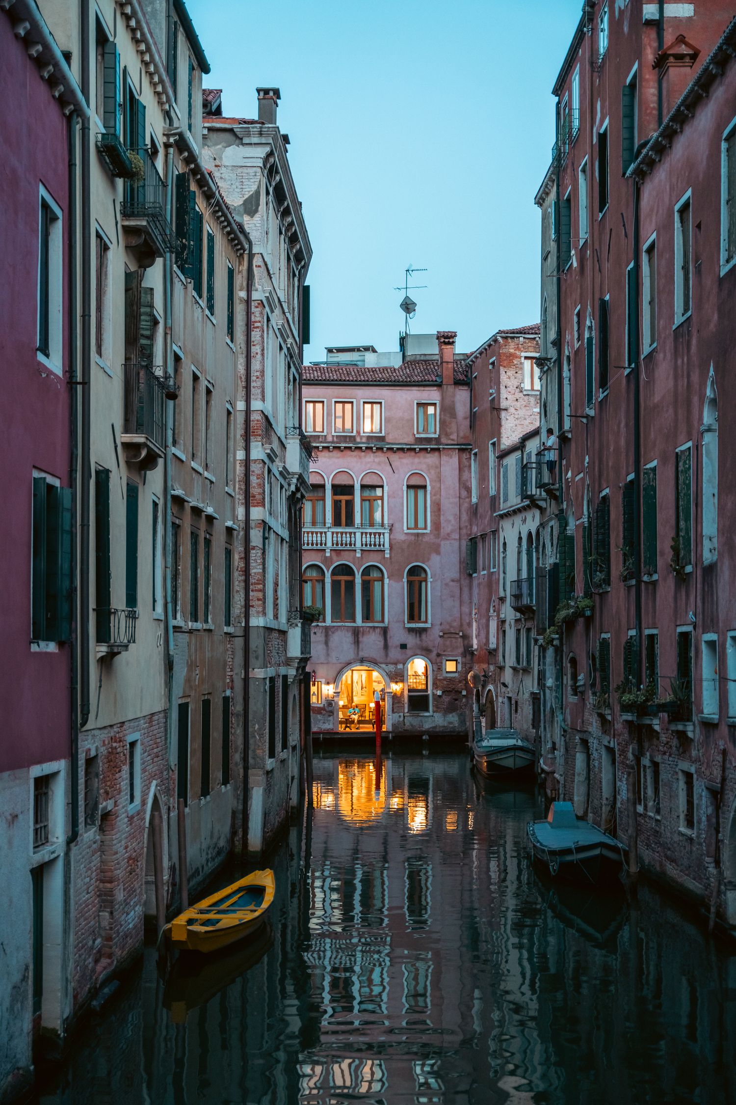 Narrow Venetian canal at dusk with warm glowing lights reflecting off water between historic buildings.
