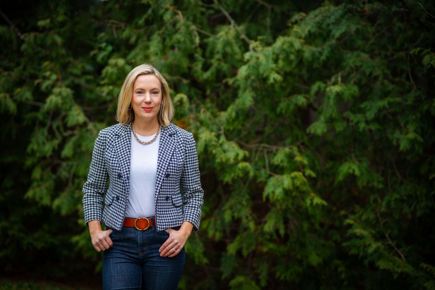 A person in a checkered blazer and jeans poses against a green leafy background outdoors.