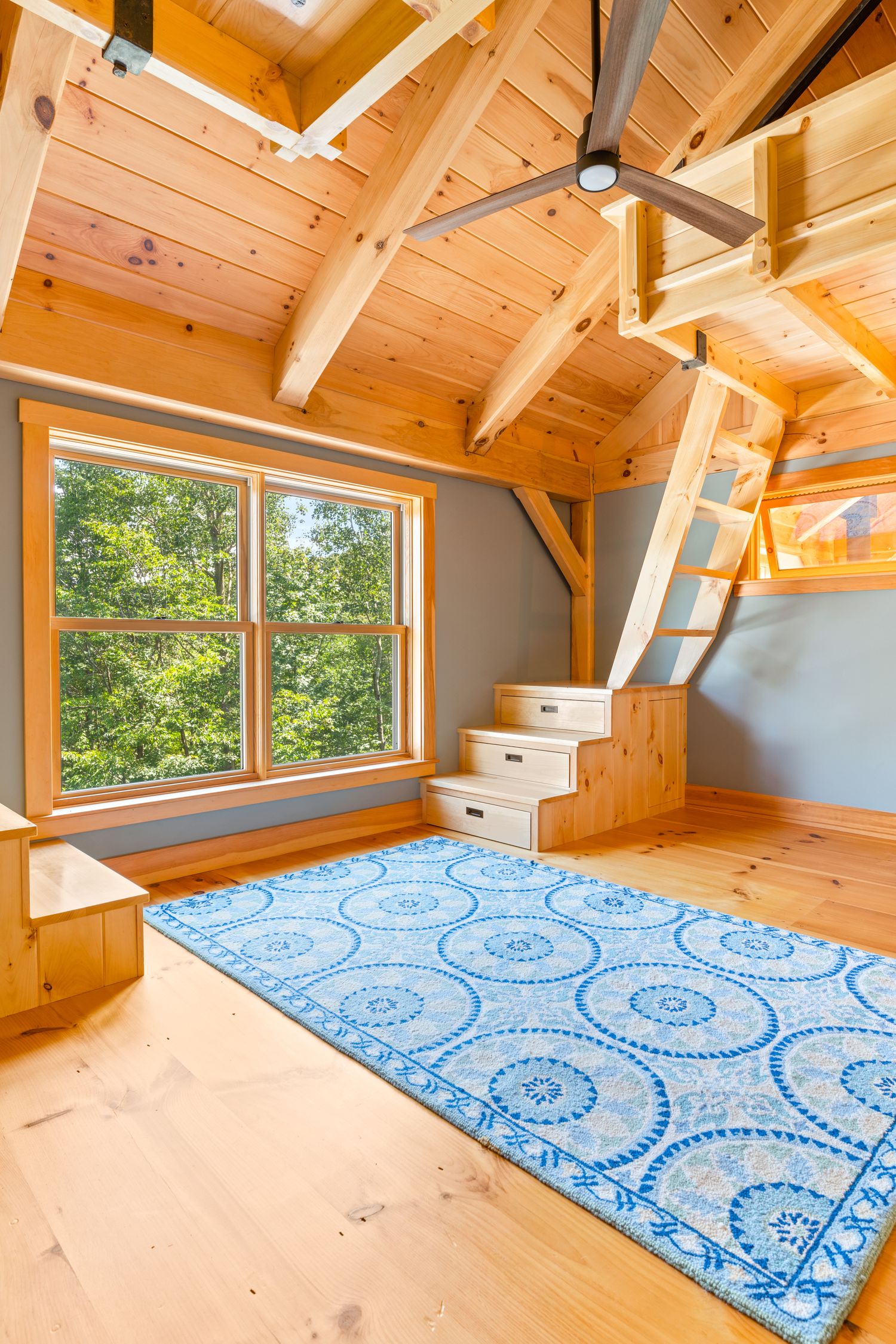 Cozy loft space with wooden beams, blue patterned rug, and large windows offering forest views.