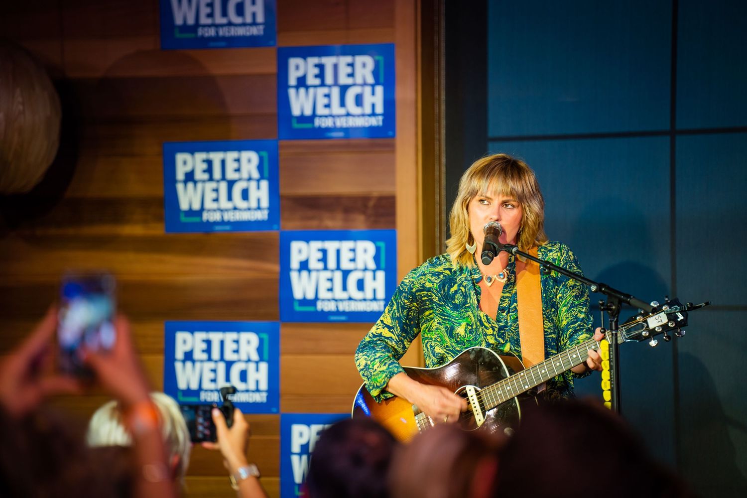 A musician performs on stage with a guitar in front of blue campaign signage and banners.