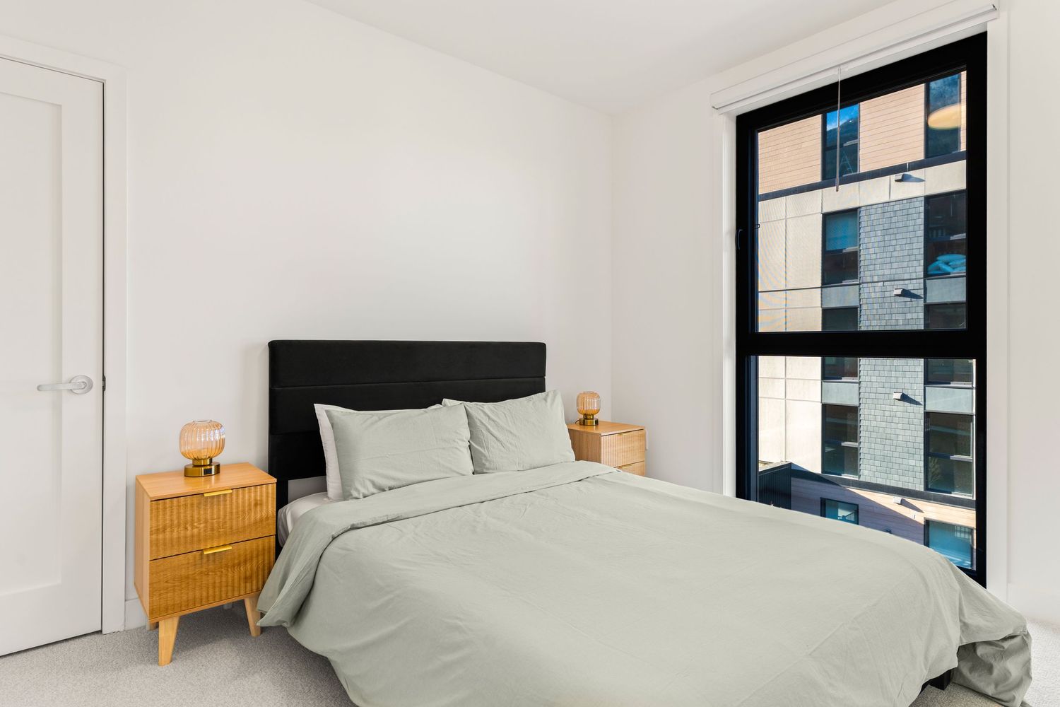 Modern bedroom interior with white walls, light gray bedding, wooden nightstand, and large windows with city views.