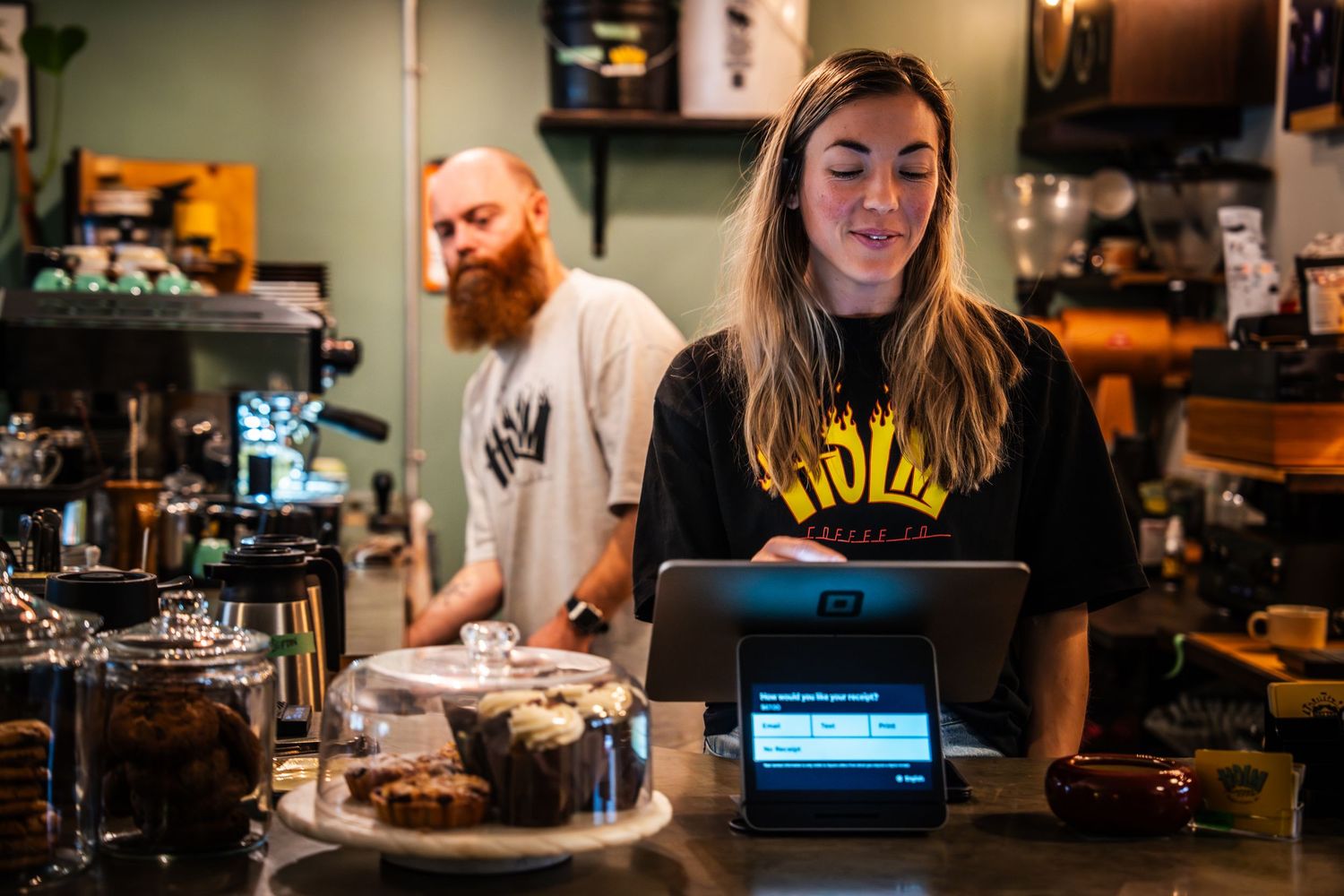 People working and socializing in a dimly lit coffee shop with laptops and beverages on wooden tables.