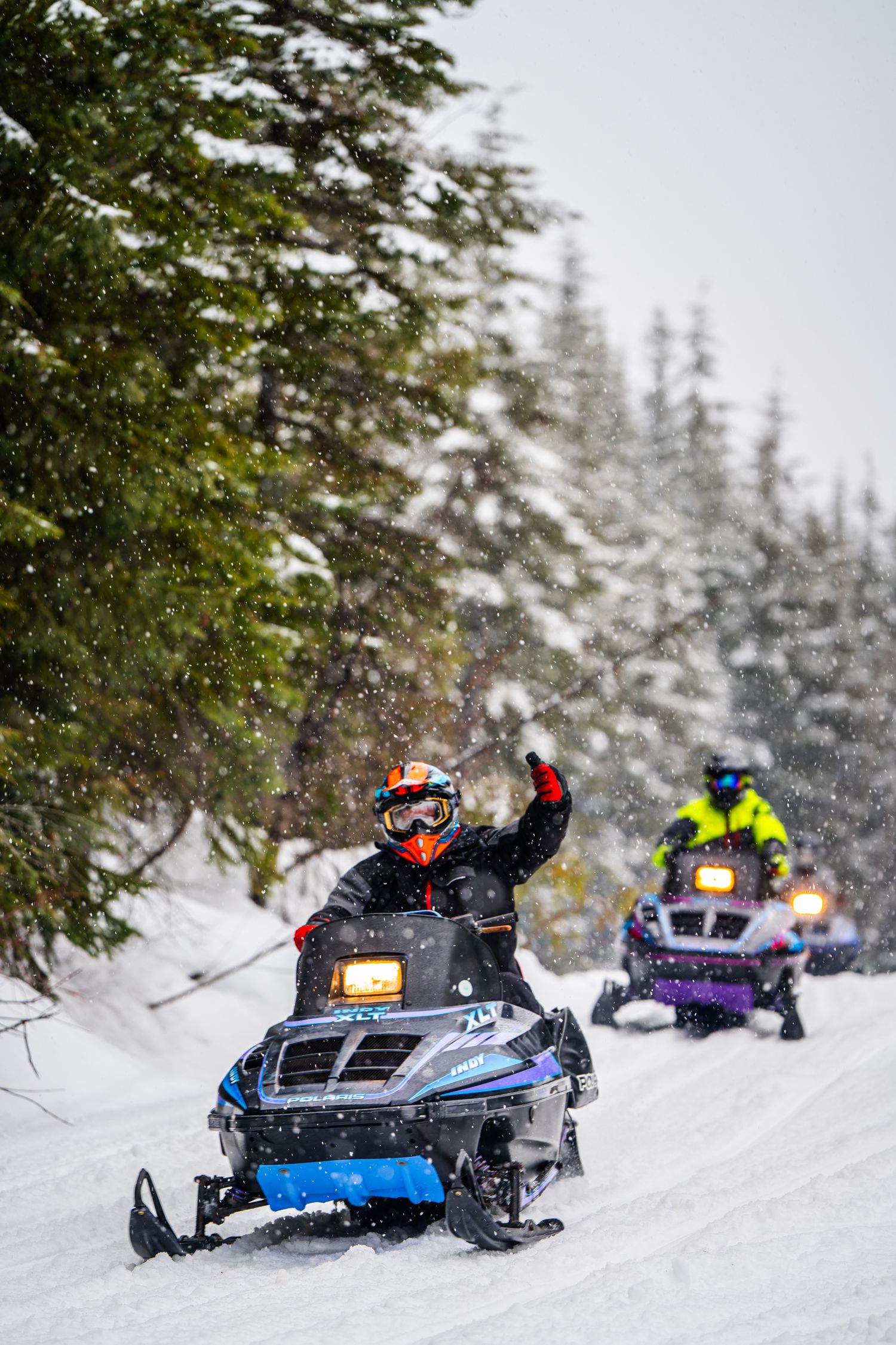 Snowmobilers riding through a snowy forest trail with evergreen trees and fresh powder in winter conditions.