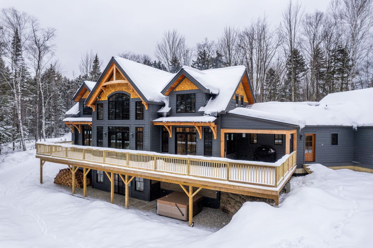 Modern mountain home exterior with snow-covered wraparound deck and dramatic timber frame architecture.