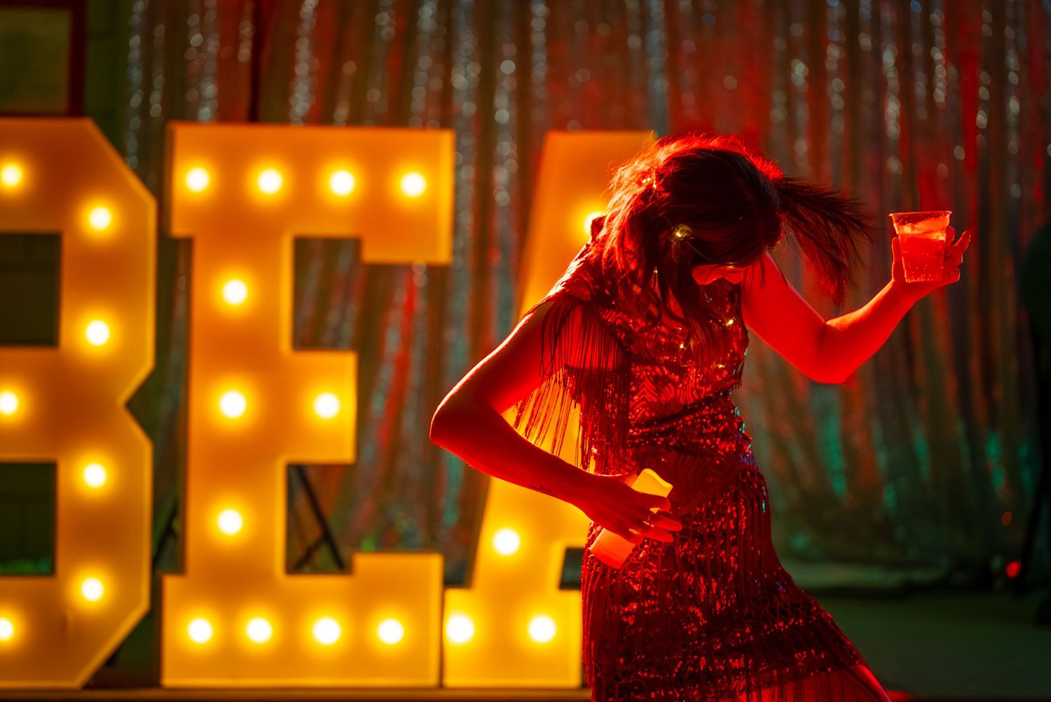 A silhouette in a red dress dances against illuminated BE letters and a forest backdrop.