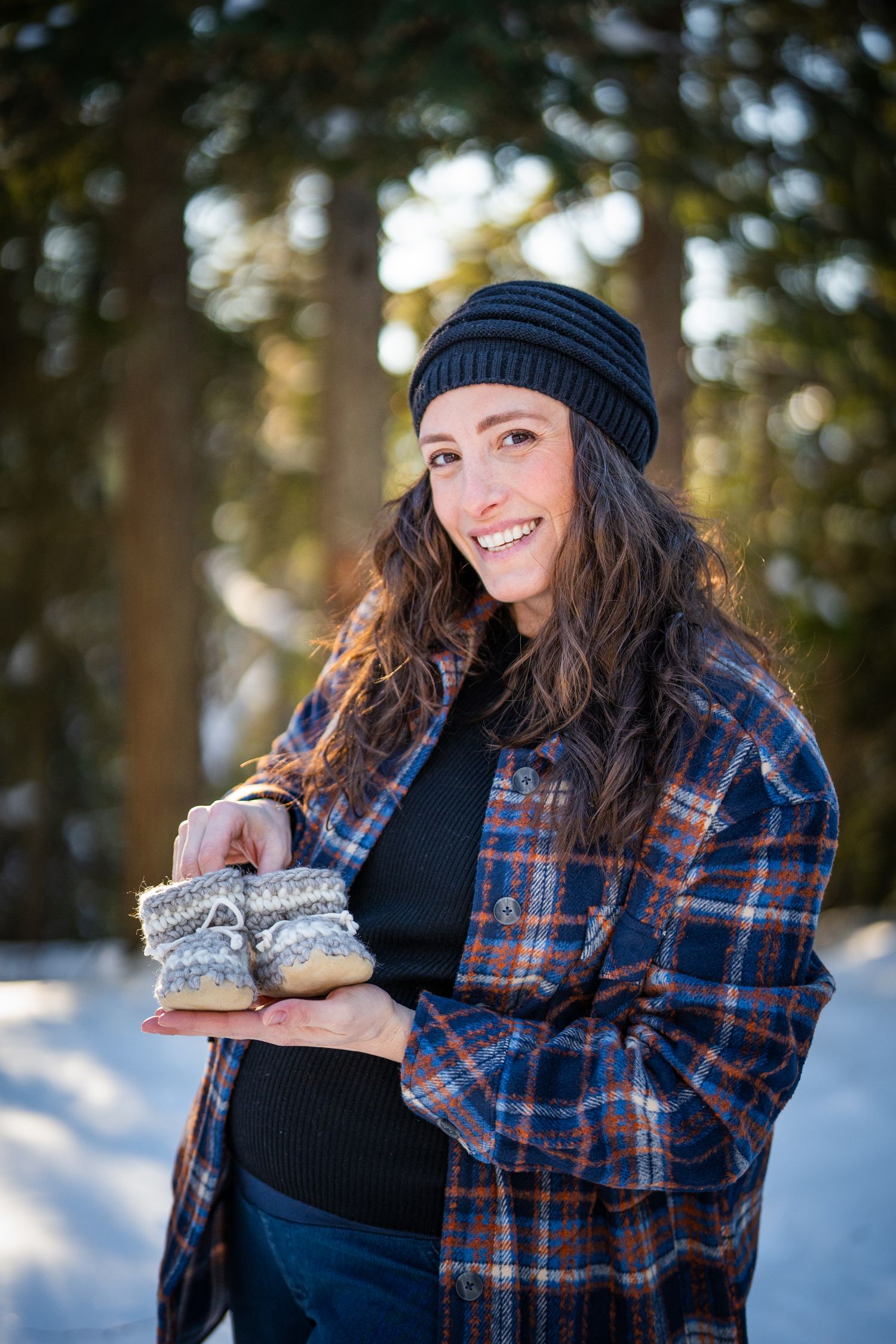 Person in winter attire holding boots in snowy forest setting with plaid flannel and knit beanie.